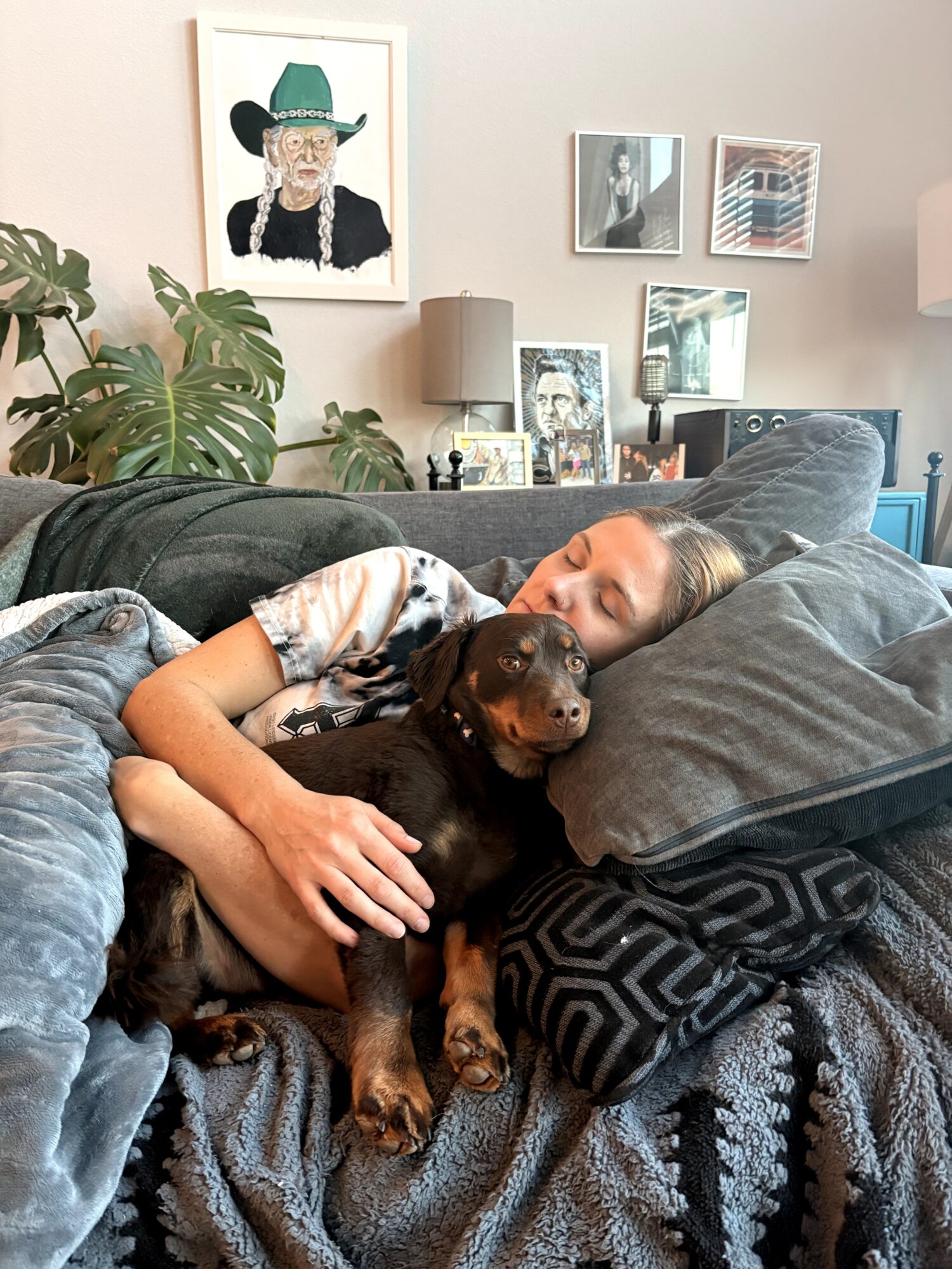 A person reclines on a couch holding a small dog, with framed pictures and a leafy plant nearby.