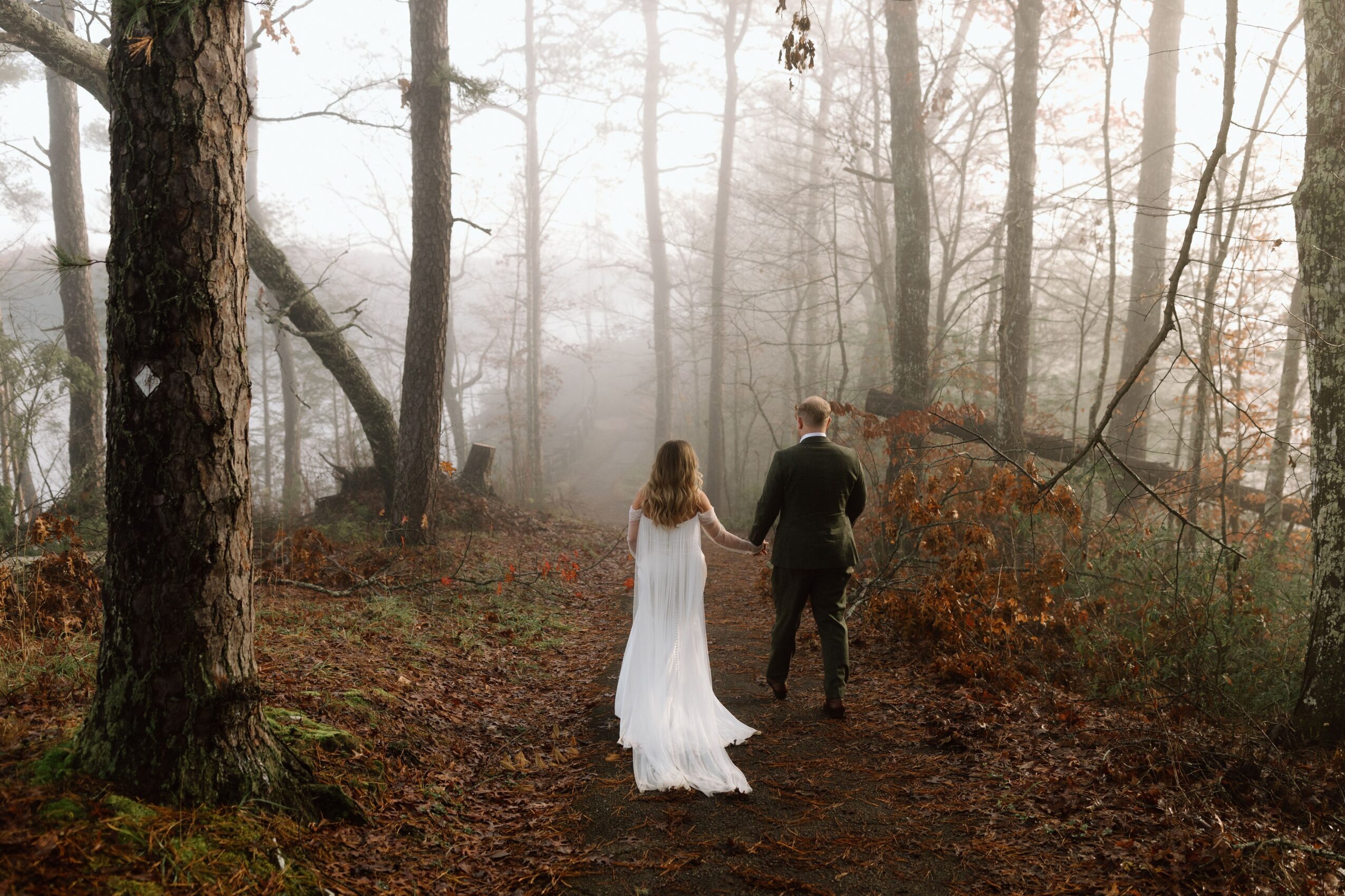 A person in a white dress walks beside another person along a foggy woodland path.
