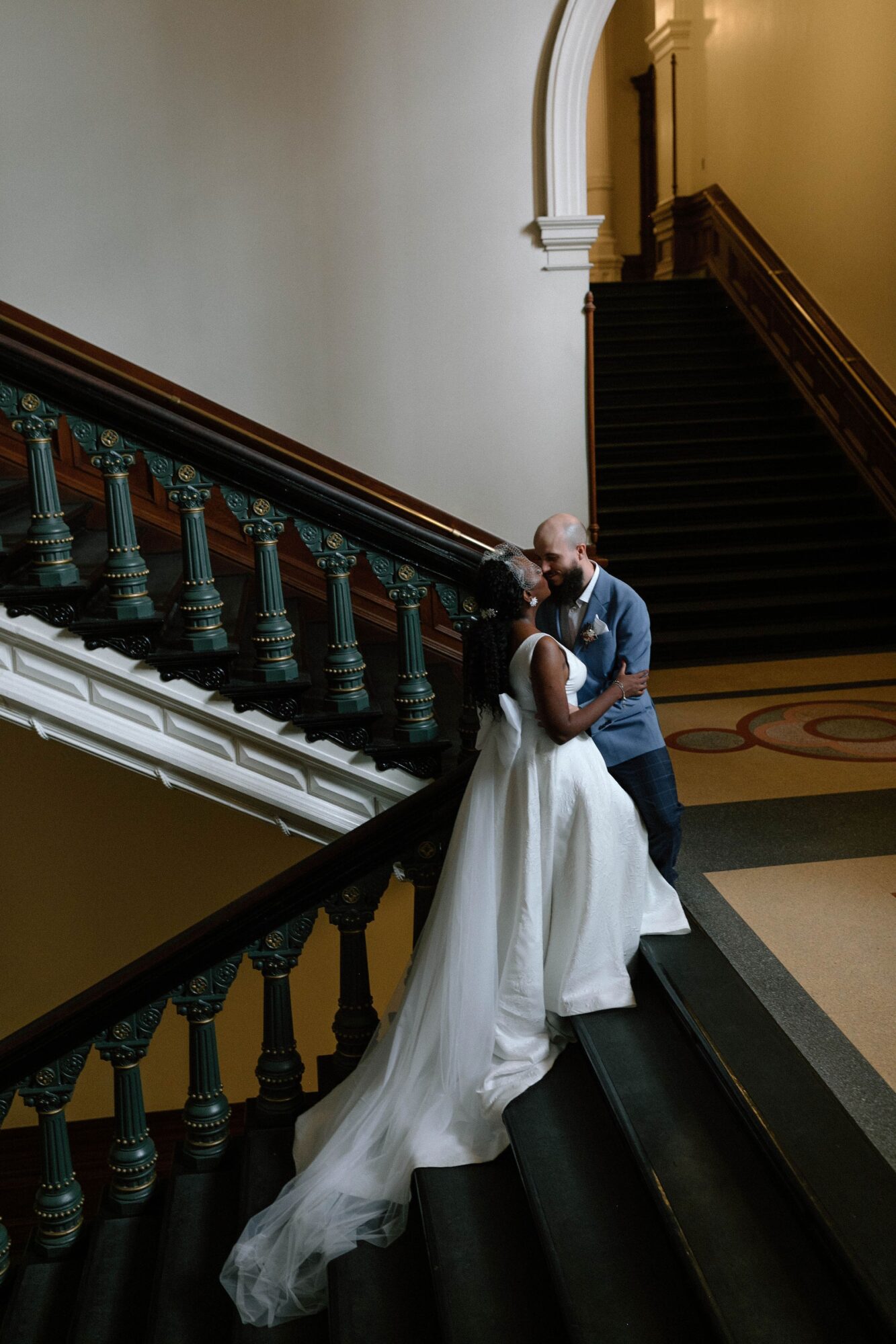 A couple embraces on a grand staircase with dark wooden railings and a long white dress train.