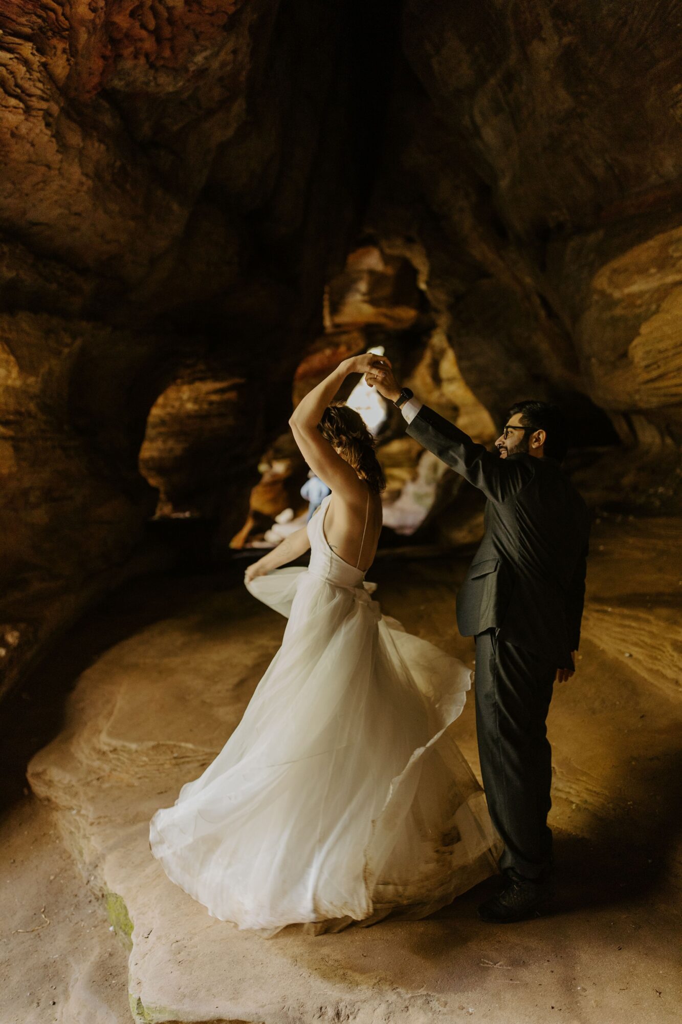A person in a white gown dances with a person in a dark suit inside a rocky cave.
