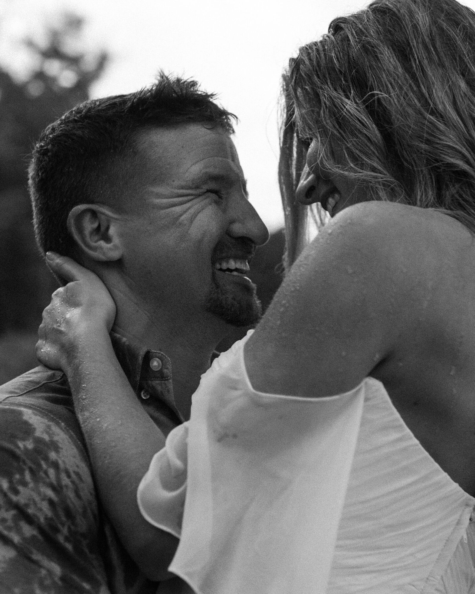 A couple embraces outdoors, smiling face to face, in a black-and-white photograph.