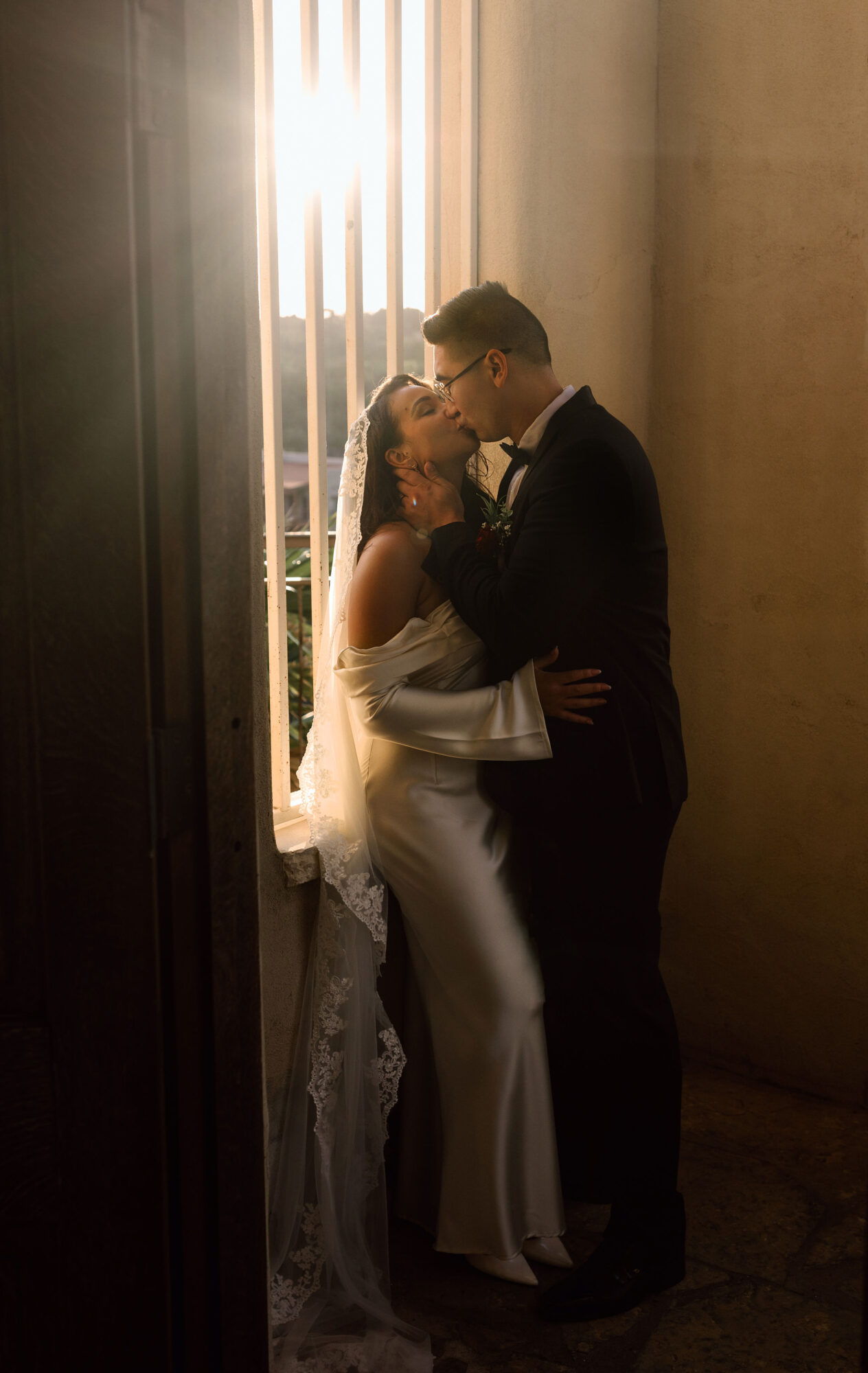 A couple embraces beside a tall, brightly lit window in a narrow hallway.