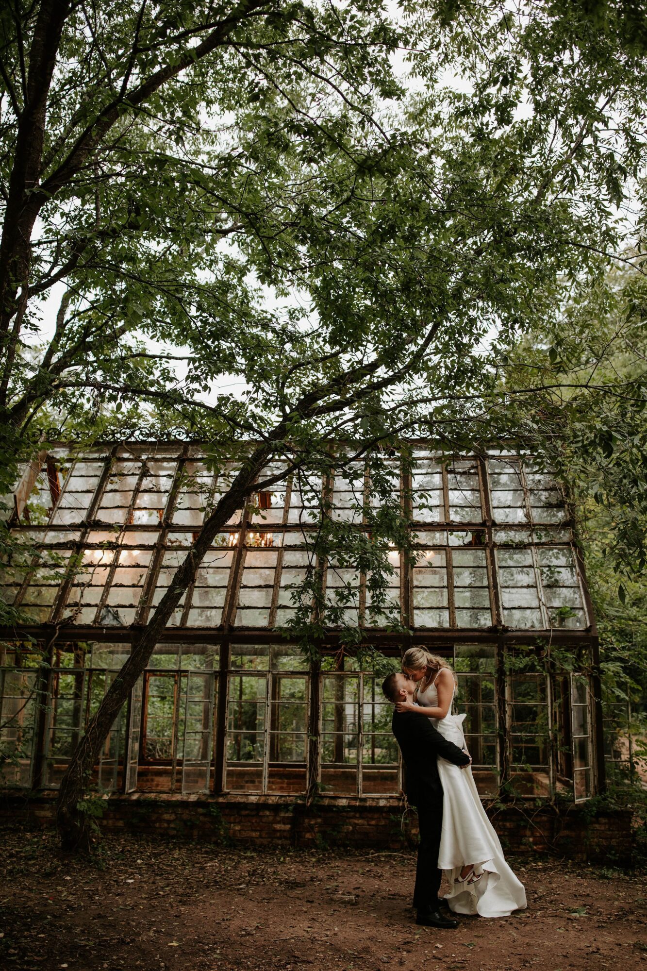 A couple embraces beneath trees in front of a glass greenhouse.