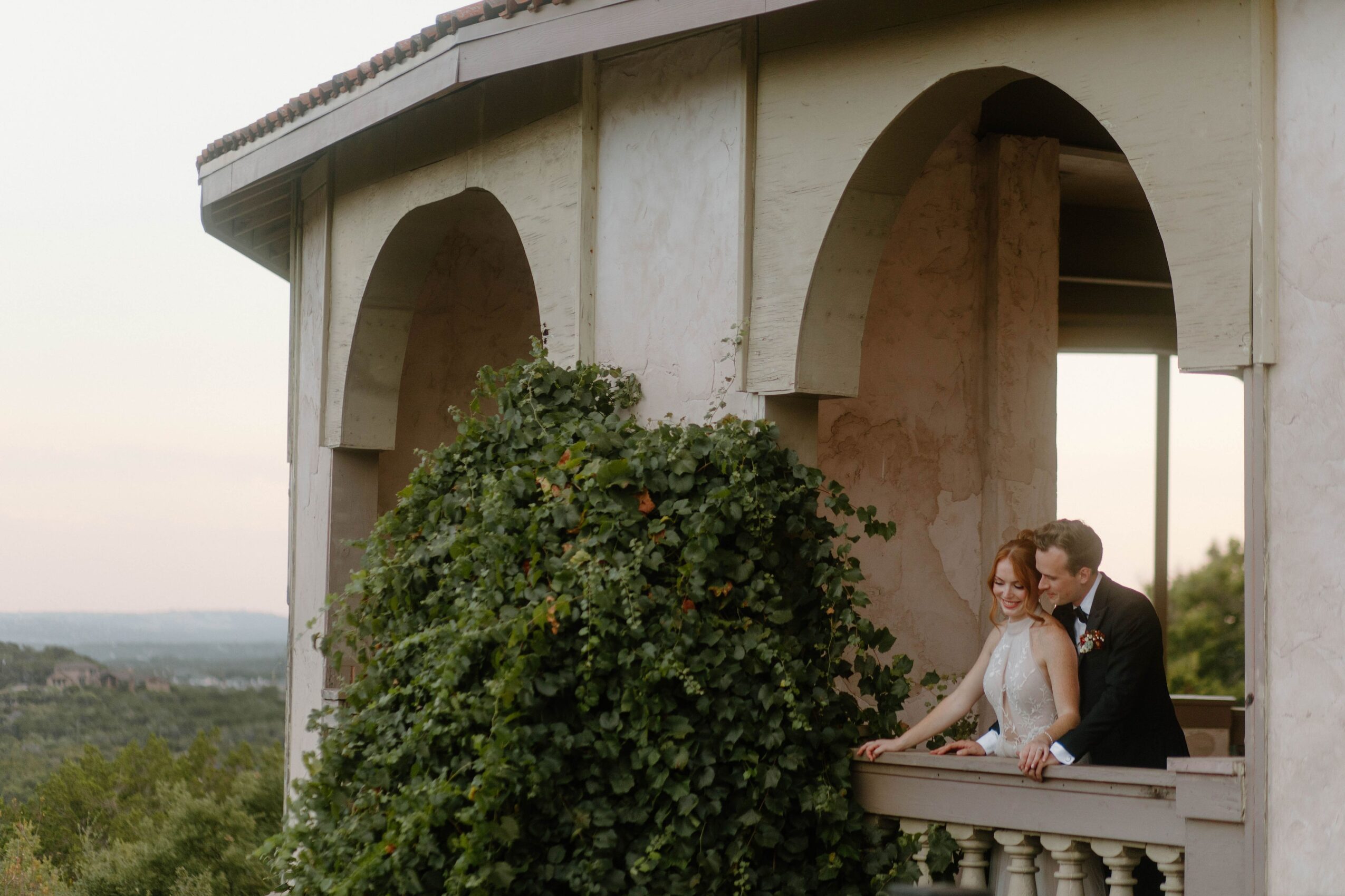 Two people stand on a balcony beside a leafy vine-covered wall with large arches and a distant landscape.