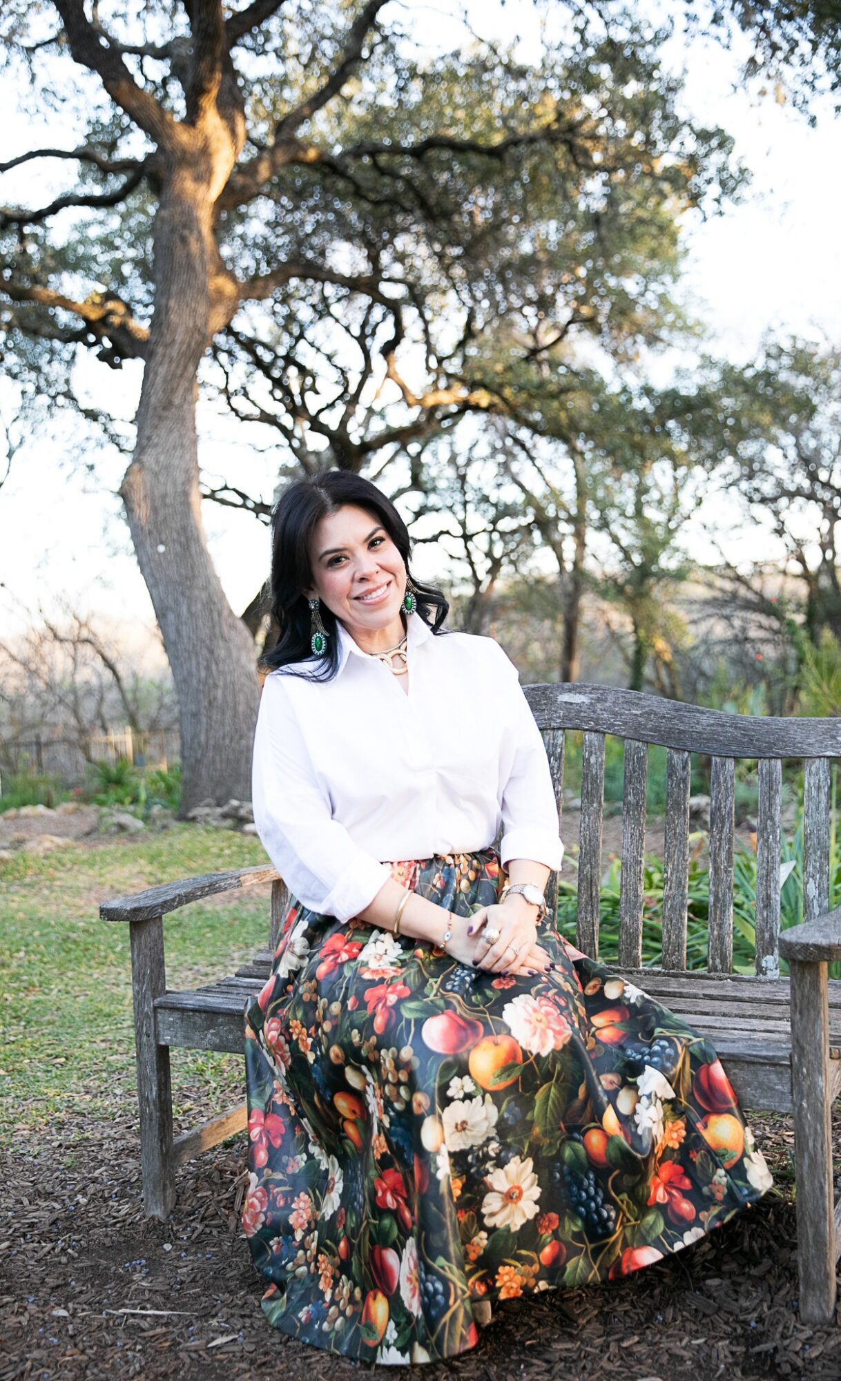 A person in a white blouse and floral skirt sits on a wooden bench beneath a large tree.
