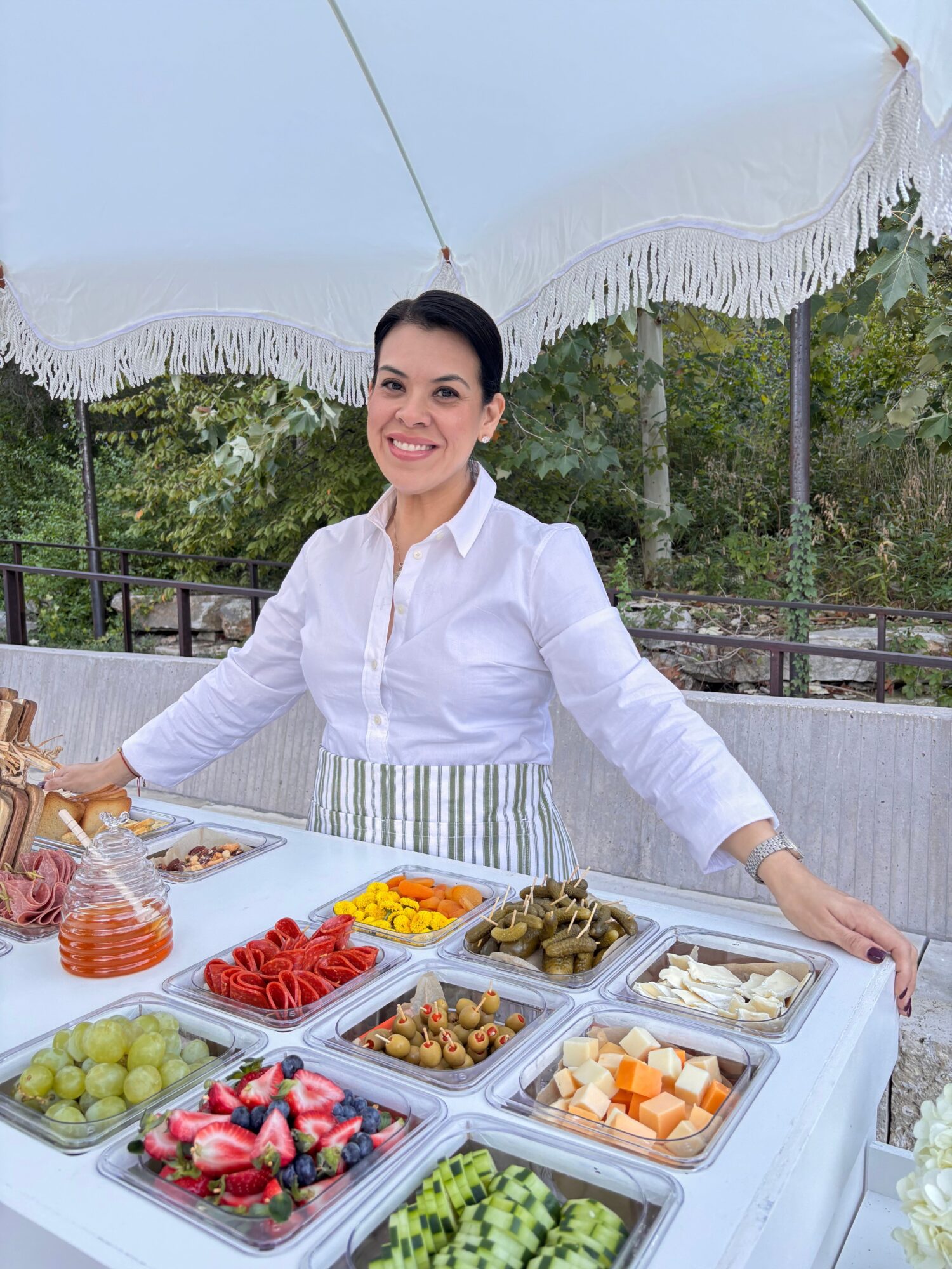 Person in a white shirt beside open trays of prepared foods under a patio umbrella.