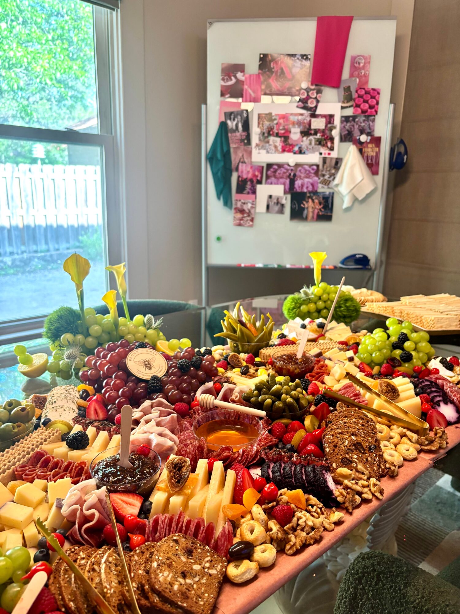 A large spread of cheeses, fruit, crackers, meats, and dips on a table beside a window and photo-covered board.