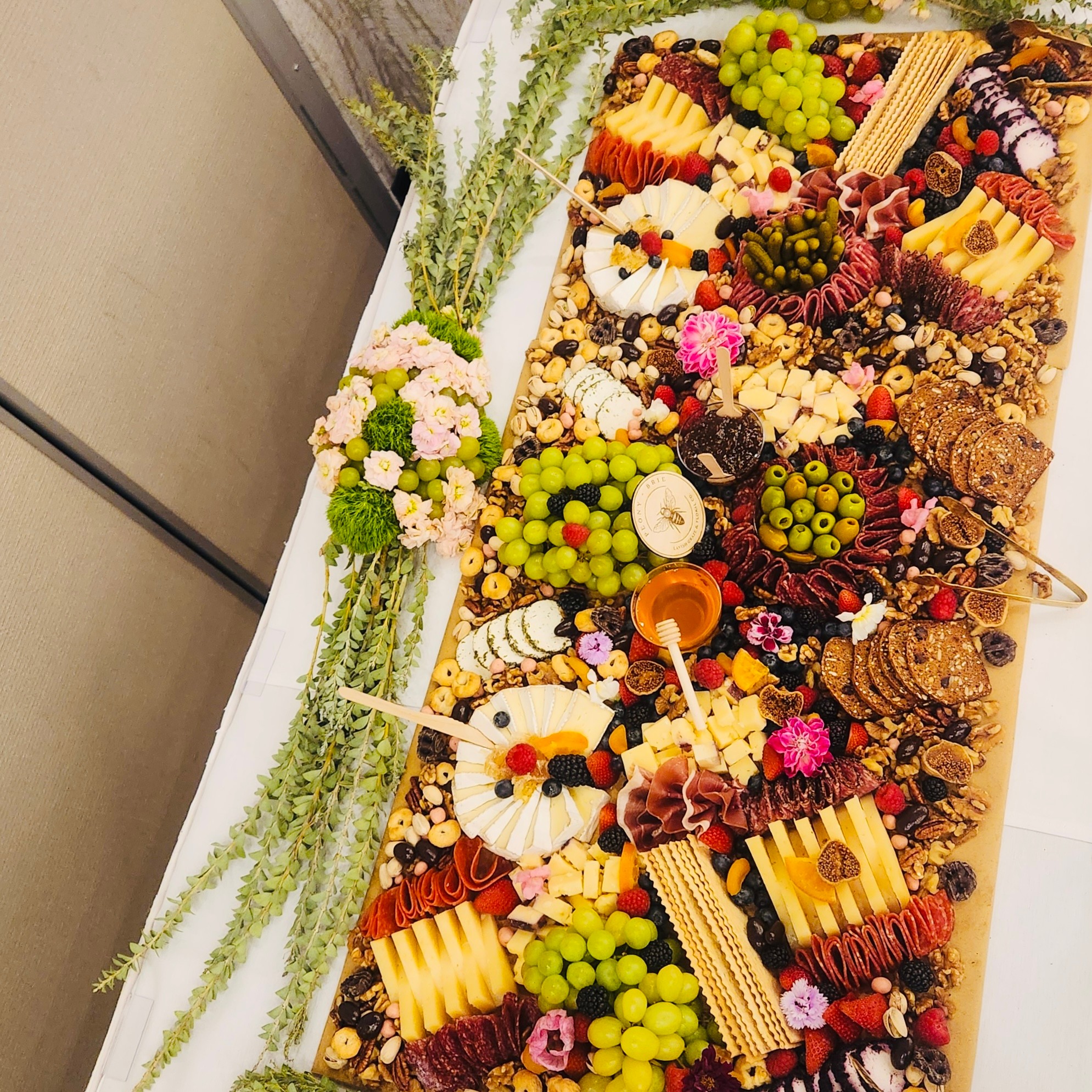 Overhead view of a long charcuterie spread with cheeses, meats, fruit, crackers, flowers, and a bowl of dip.