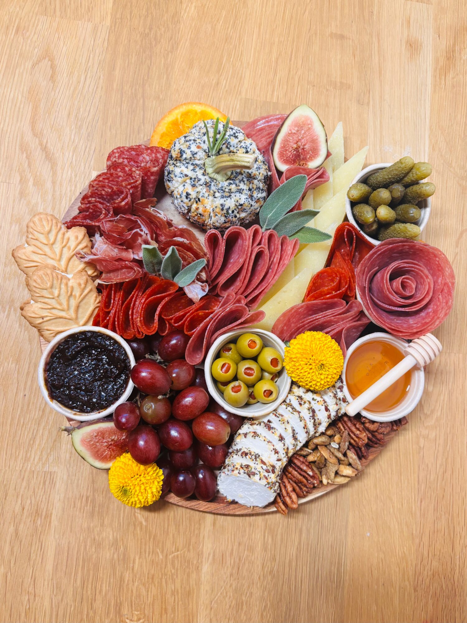 Round charcuterie platter with sliced meats, cheeses, olives, fruit, crackers, and dips on a wooden table.