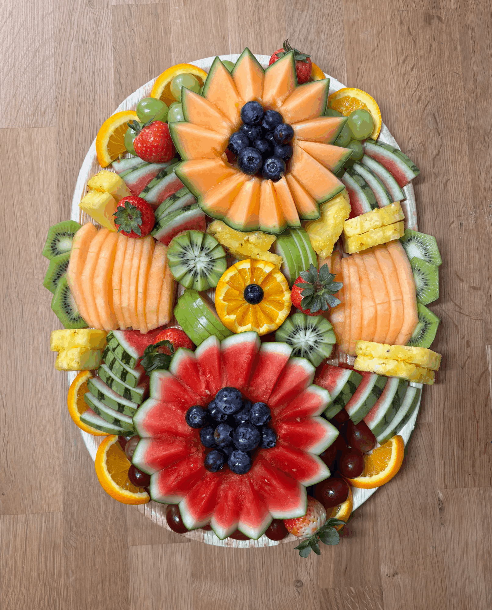 An oval platter of assorted fruit arranged in orange and red flower shapes on a wooden floor.