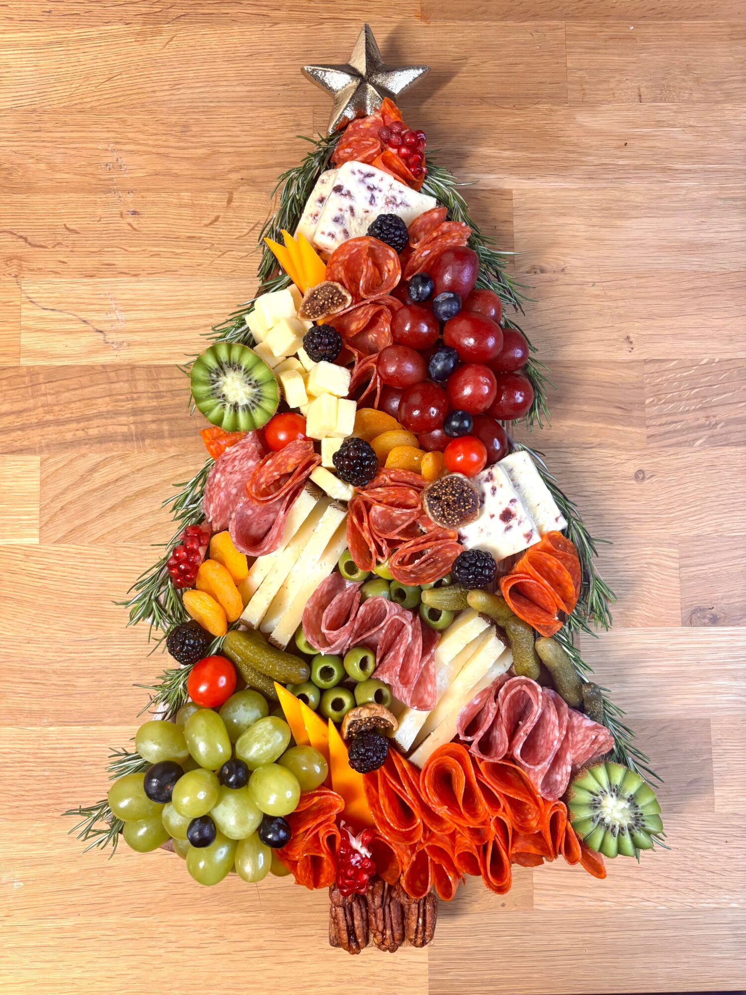 A Christmas tree-shaped arrangement of meats, cheeses, fruit, olives, and herbs on a wooden floor.