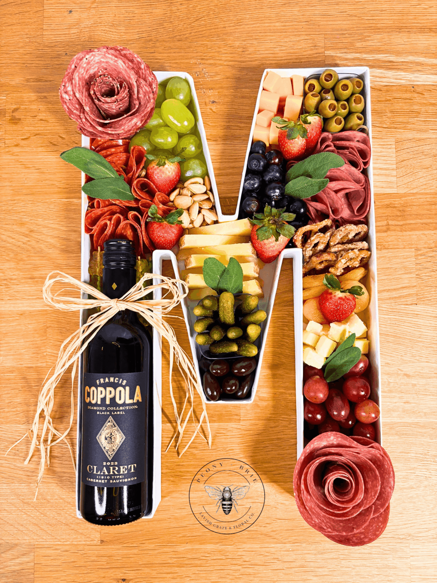 Wooden letter M filled with fruit, olives, cheese, and cured meats beside a wine bottle and rose-shaped salami.