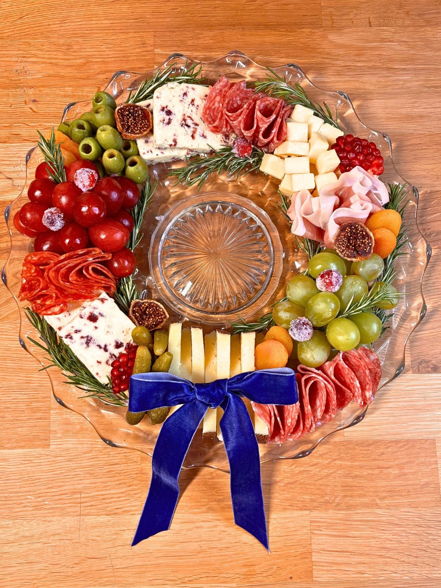 Round platter arranged with cheeses, sliced meats, olives, grapes, fruit, and a blue ribbon on a wooden table.
