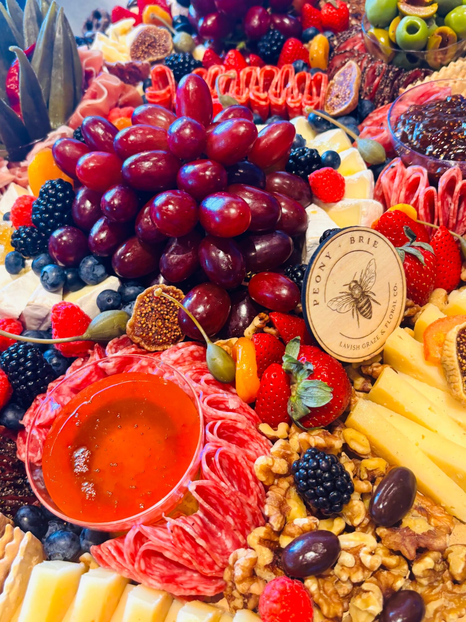 An abundant platter of grapes, berries, sliced meats, cheese, nuts, and a bowl of red sauce.