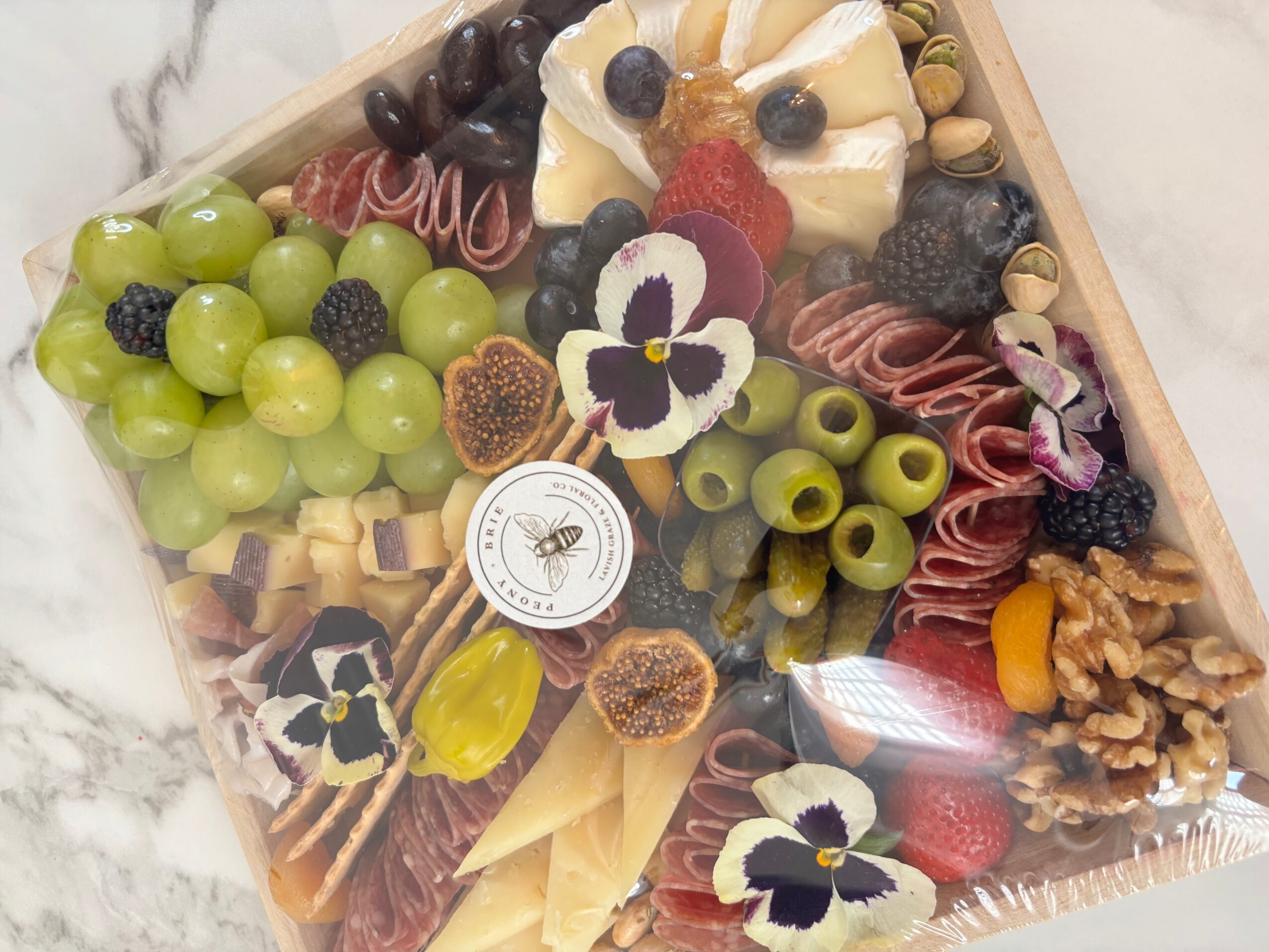 A rectangular platter arranged with grapes, olives, cheeses, cured meats, nuts, dried fruit, and decorative edible flowers.