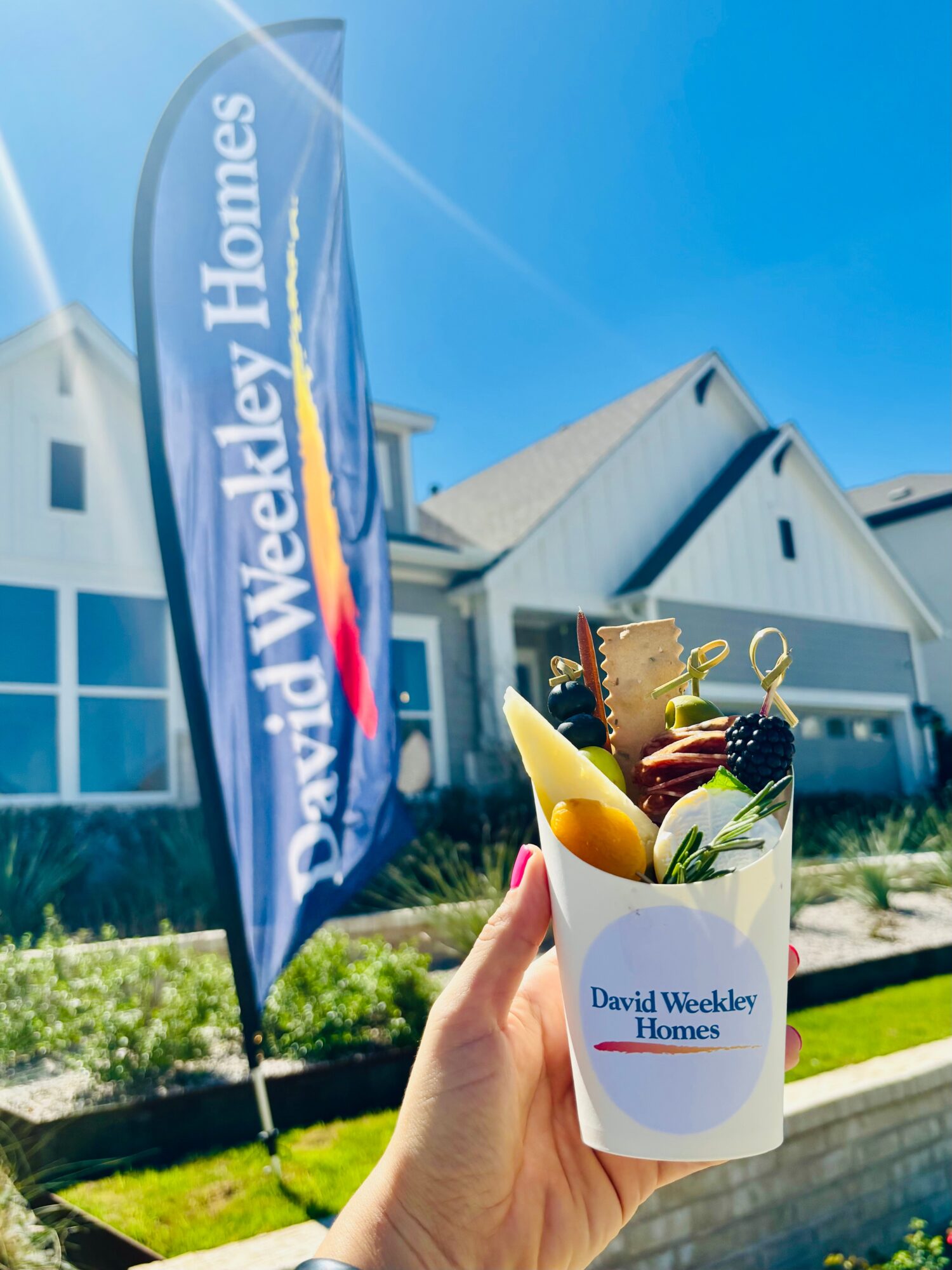 A hand holds a branded cup filled with snacks outside a house, with a real estate banner nearby.