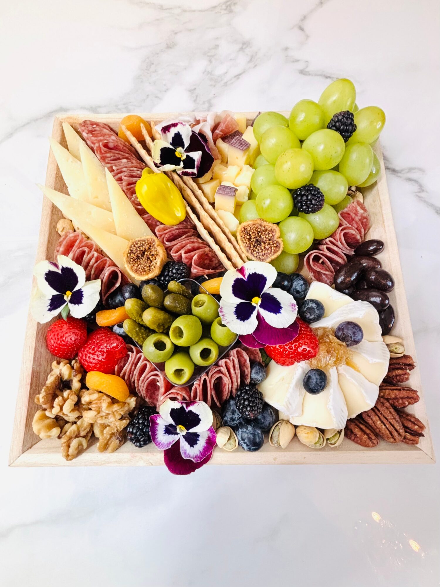 A rectangular platter of grapes, cheeses, cured meats, olives, nuts, berries, and edible flowers.