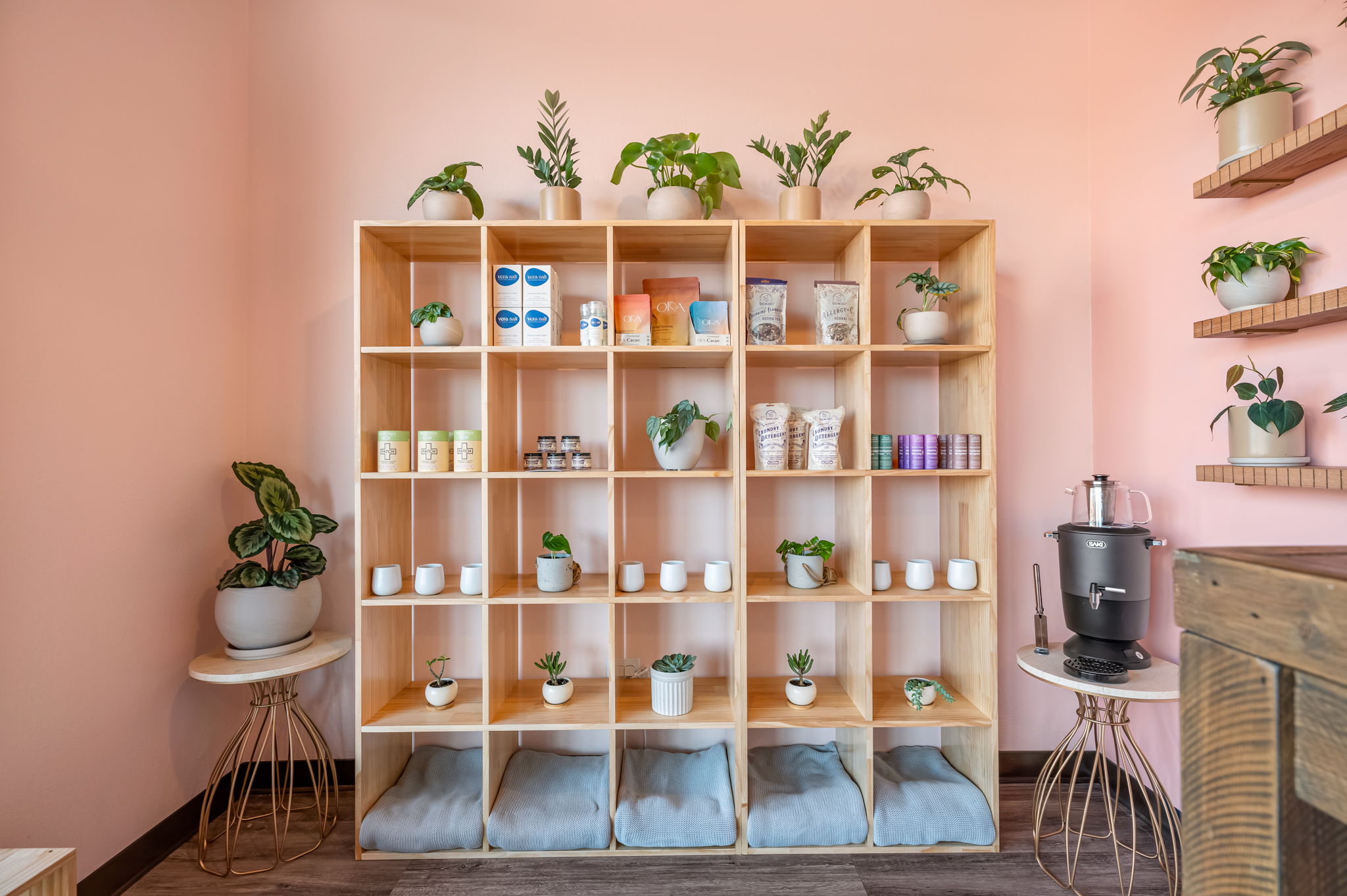 A wooden cubby shelf filled with jars and small plants stands against a pink wall, with plants on side shelves and tables.