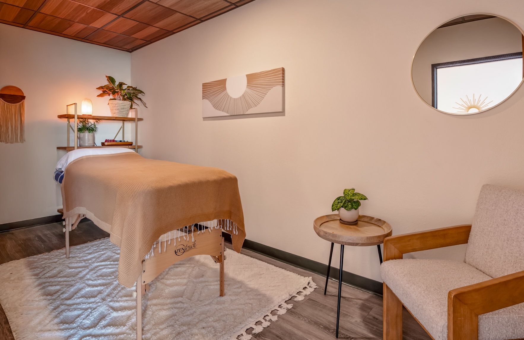 A massage table with a beige cover sits in a softly lit room beside a chair, side table, plants, and wall art.
