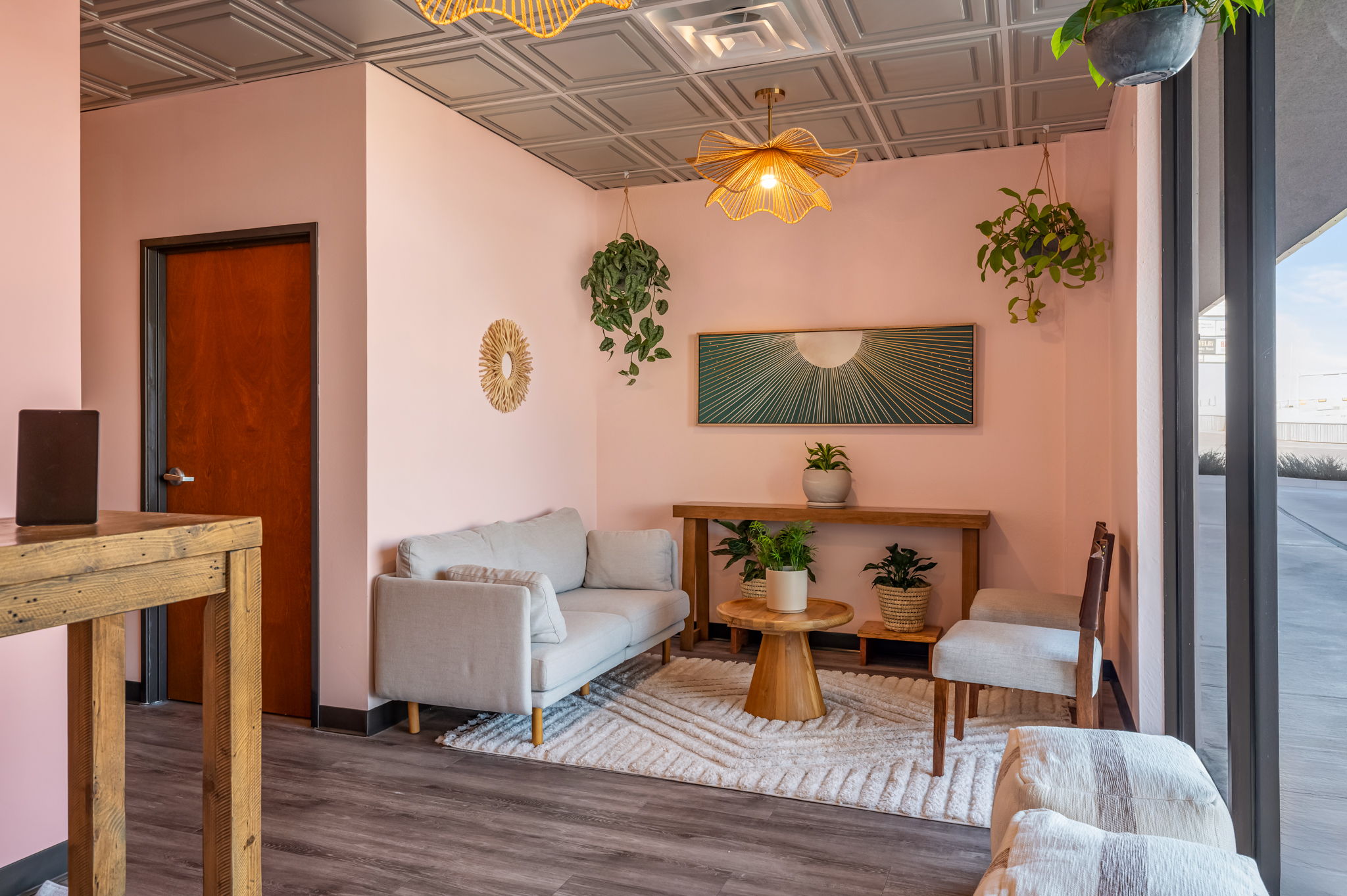 A pink living room with a sofa, chairs, wooden tables, plants, and a patterned rug beside large windows.