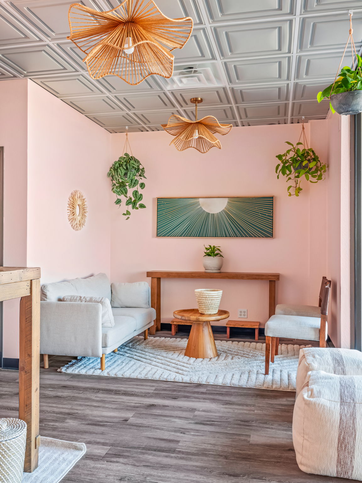 Living room with pink walls, white seating, a wooden coffee table, hanging plants, and woven pendant lights.