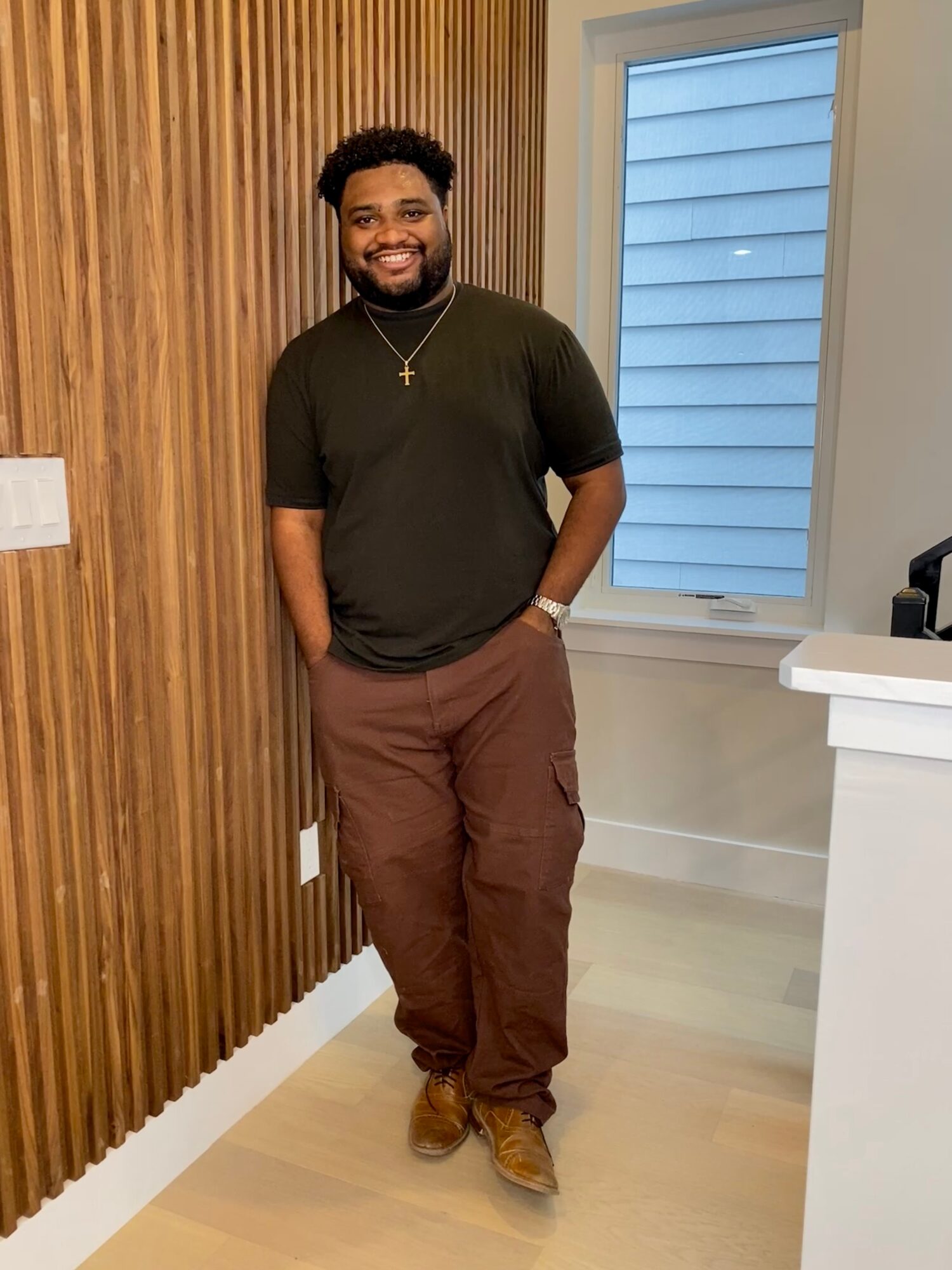 A person stands smiling beside a wood-paneled wall and a window.