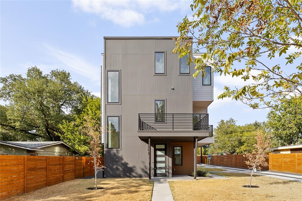 Modern three-story house with a gray facade, balconies, wooden fences, and surrounding trees.