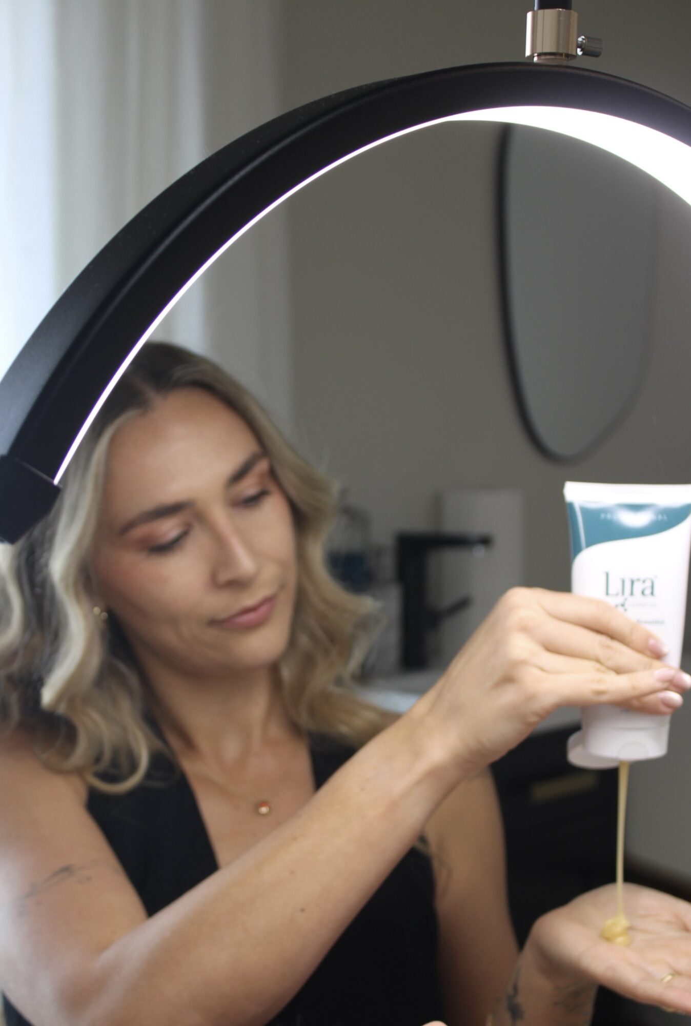 A person holds a skincare tube beneath a lit ring light while dispensing product into an open palm.