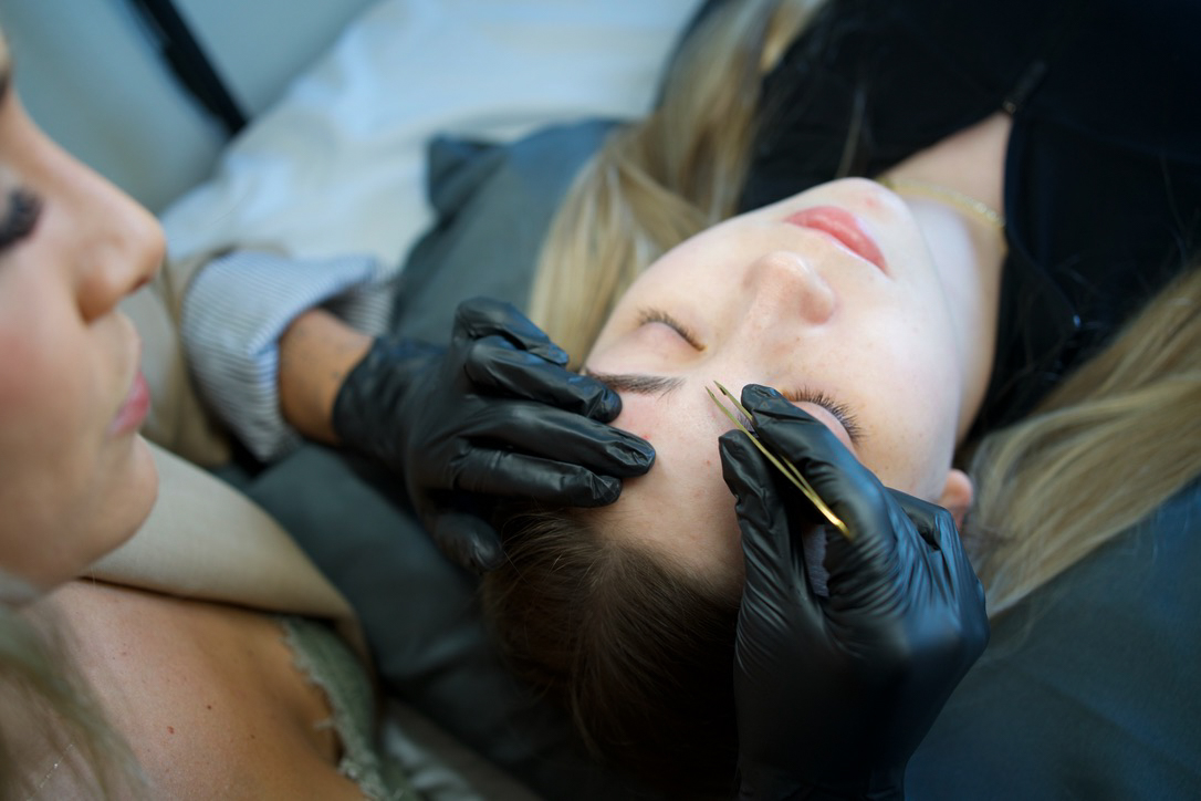 A person wearing black gloves applies a small tool near another person's eyebrow as they lie back.