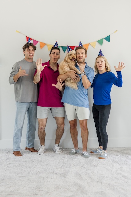 Five people wearing party hats stand together, with one person holding a small dog and colorful pennants hanging behind them.