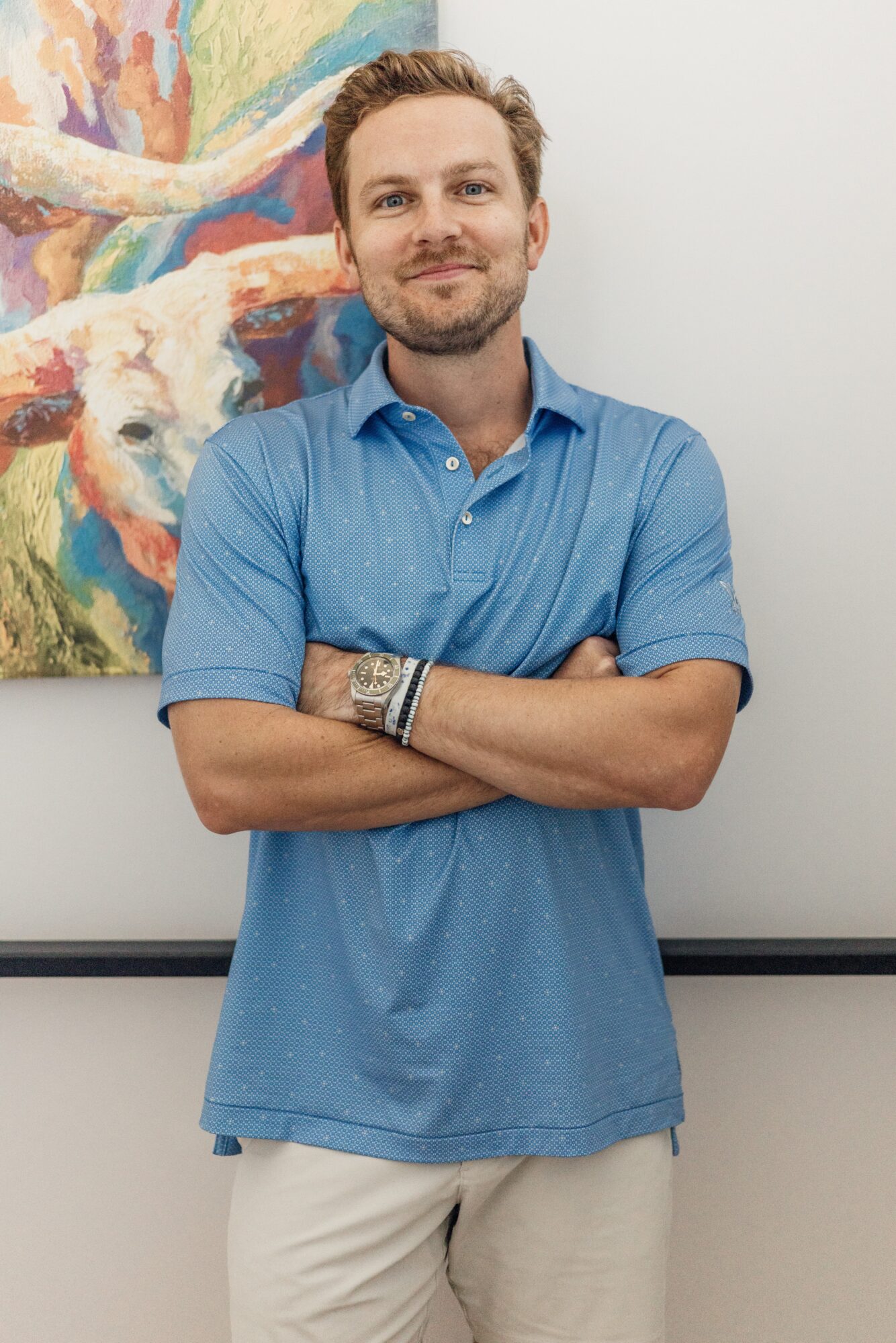 A person in a blue polo shirt and light shorts stands with arms crossed in front of a colorful painting.