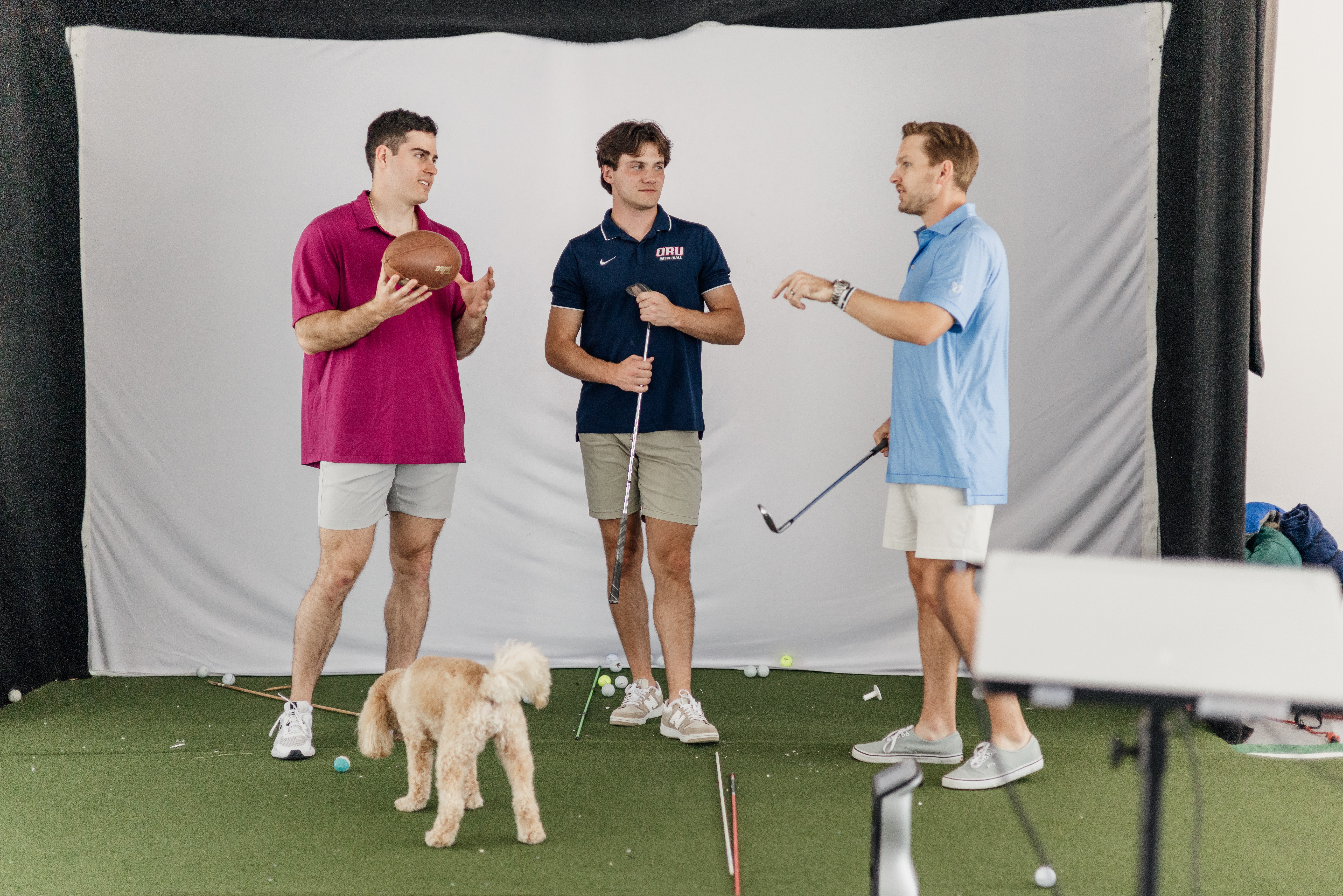 Three people stand on an indoor turf area with a dog, golf clubs, and a ball.