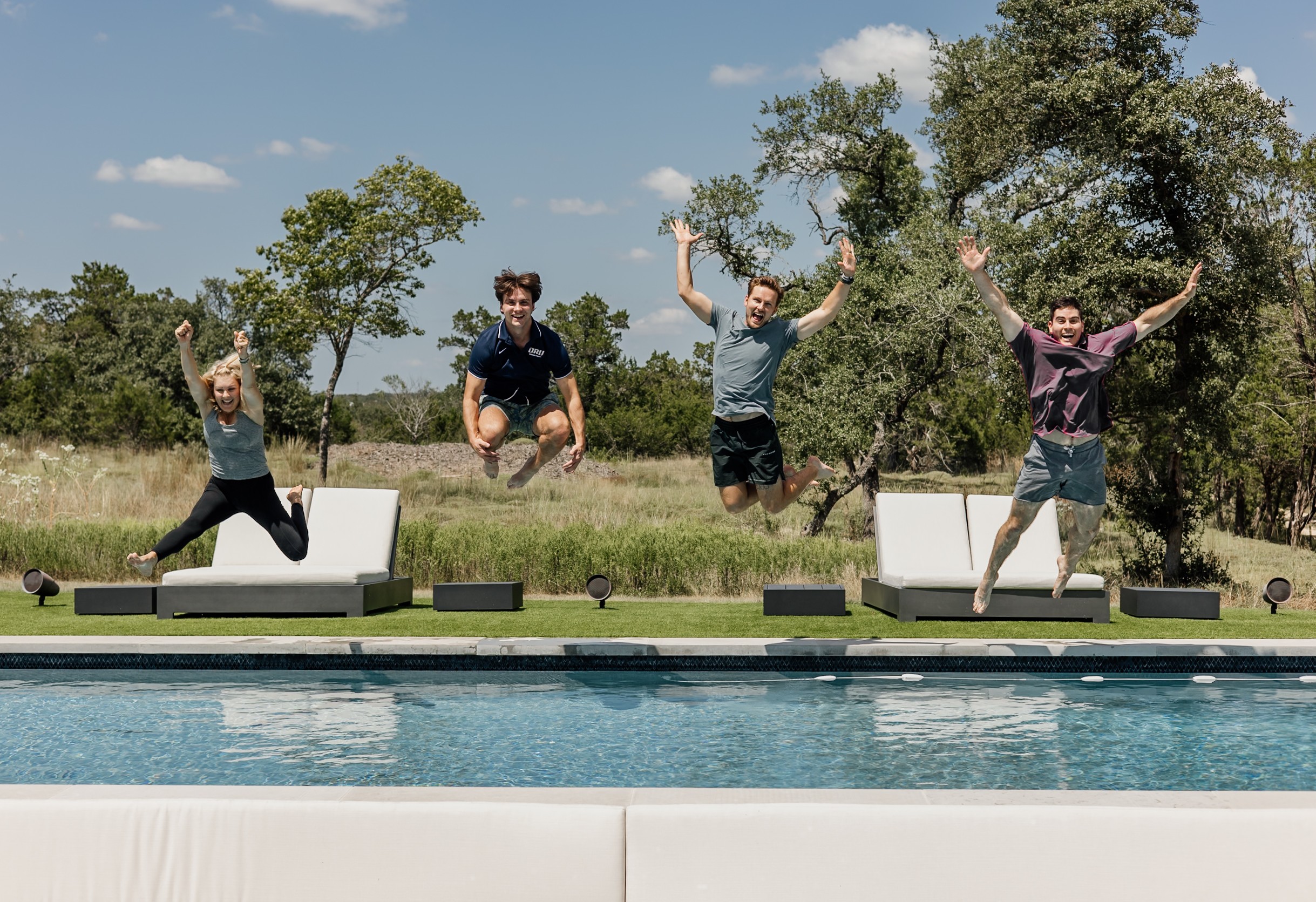 Four people jump beside a swimming pool, with trees and grass in the background.