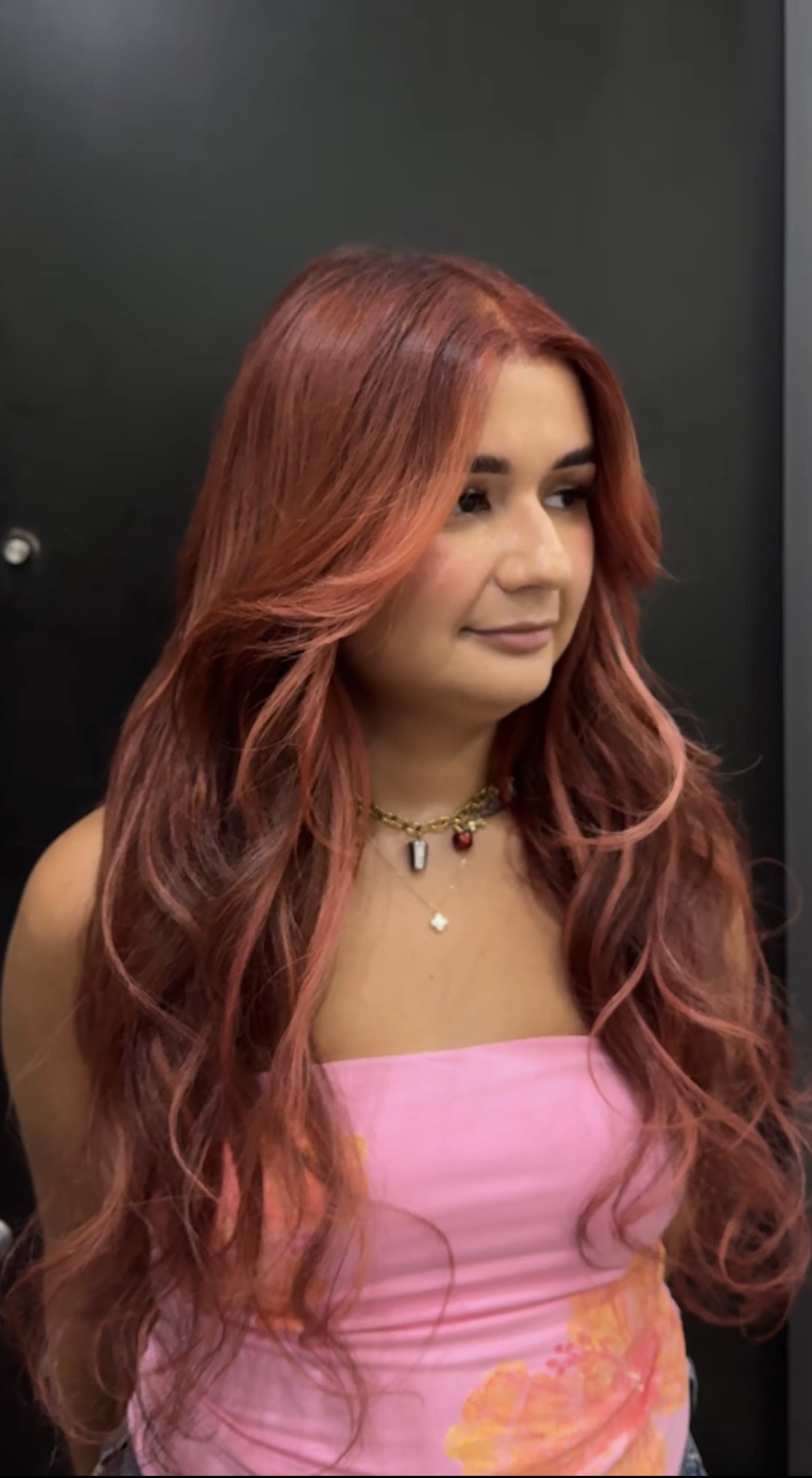 Person with long reddish hair wearing a pink strapless top, standing against a dark background.