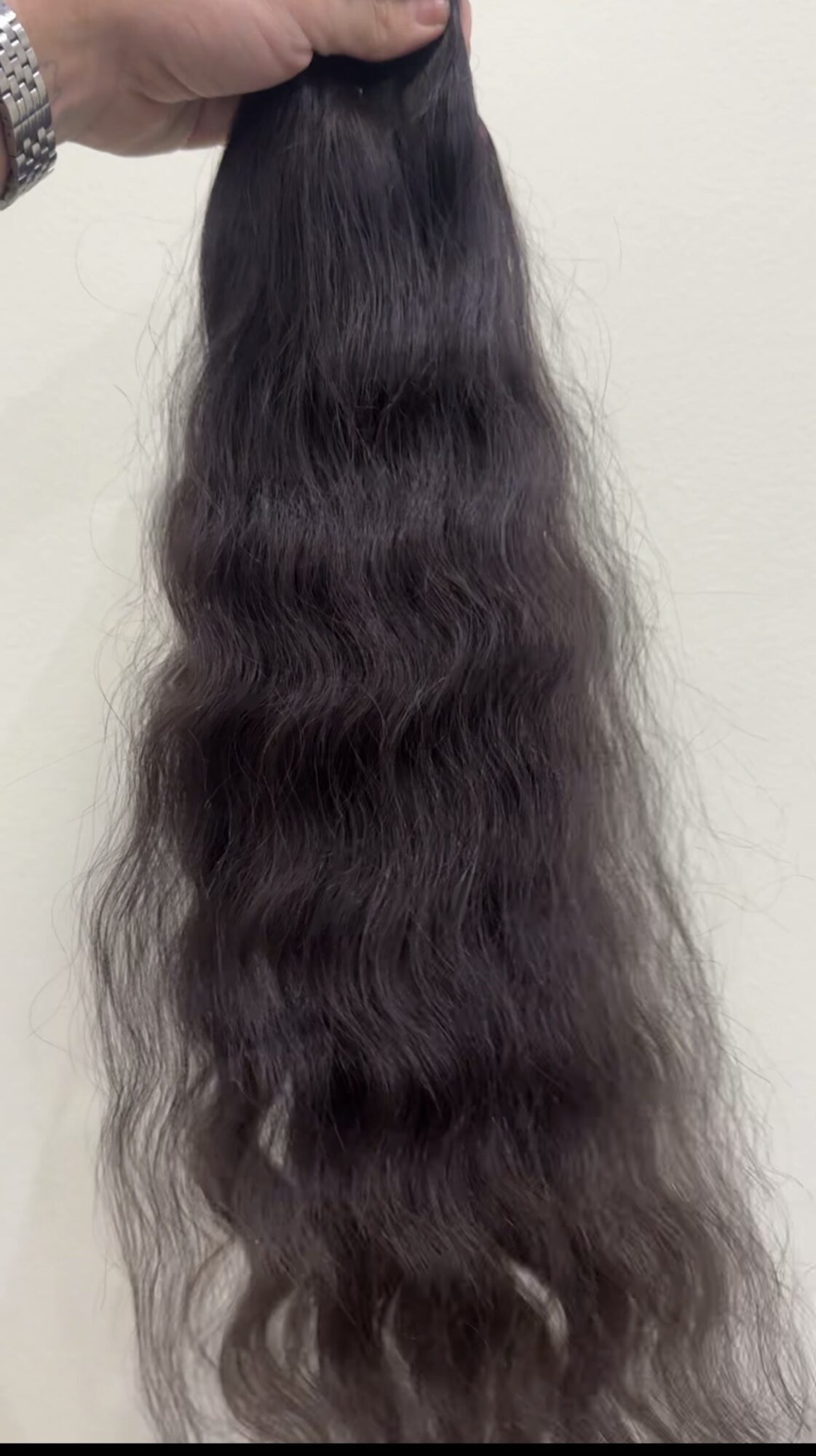 A hand holds a long, dark wavy hairpiece against a light-colored wall.