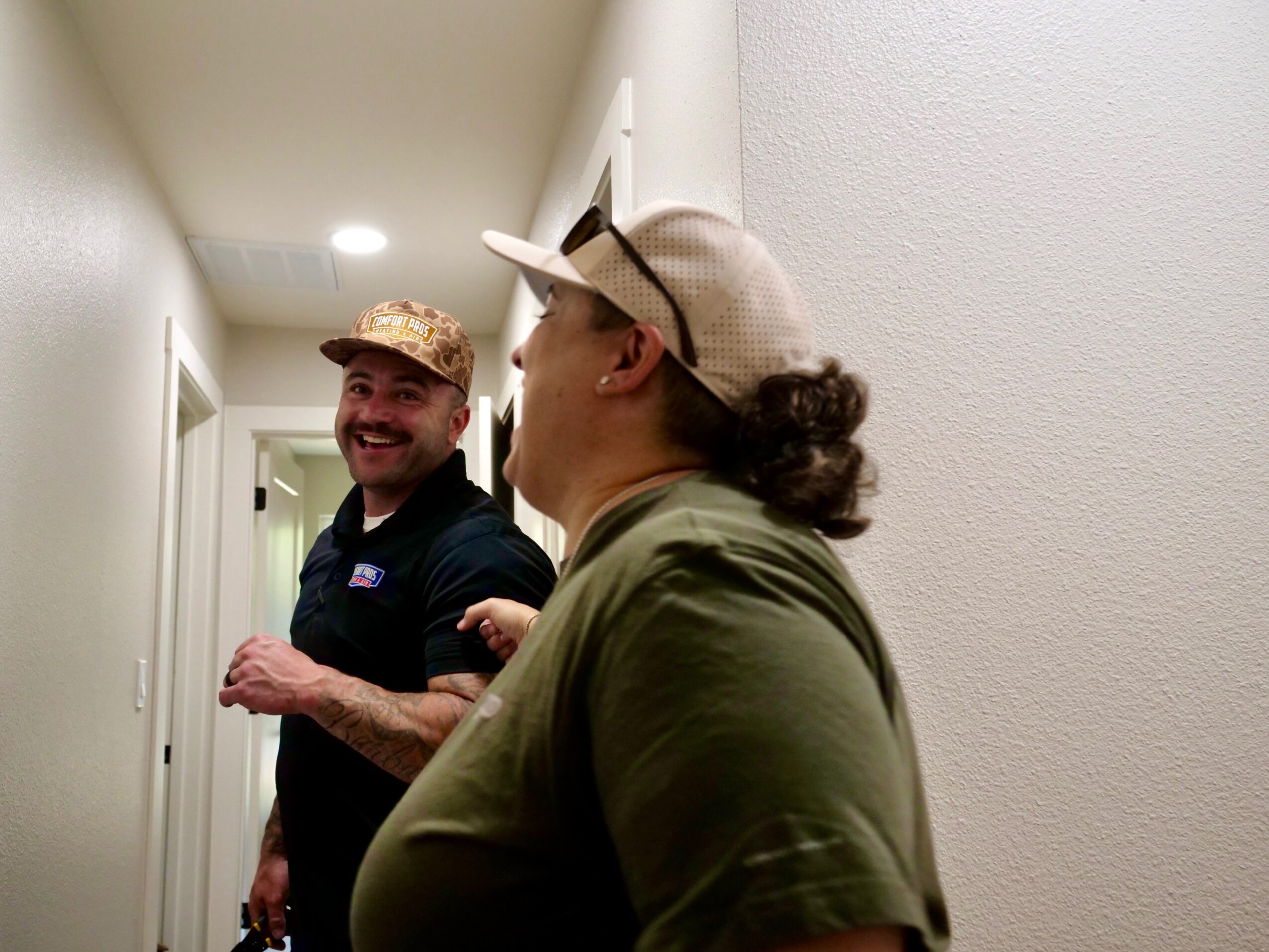Two people stand in a narrow hallway, one wearing a cap and black shirt, the other wearing a light cap and green shirt.