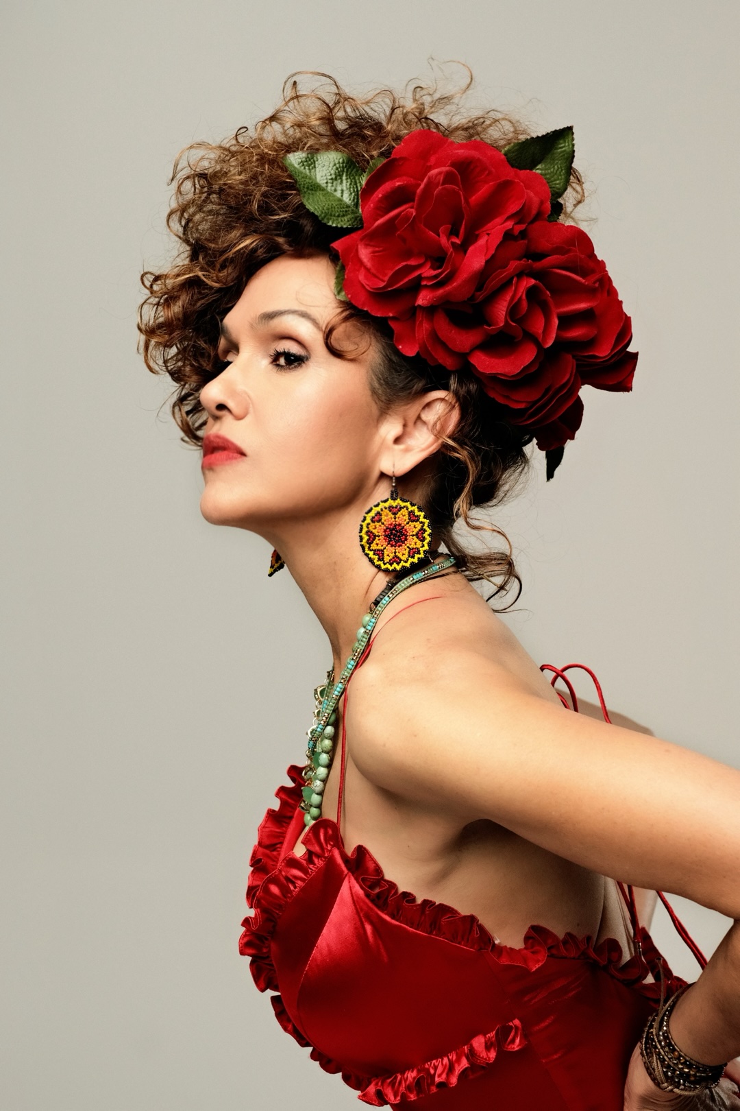 Profile portrait of a person wearing a large red flower headpiece, colorful earrings, and a red ruffled dress.