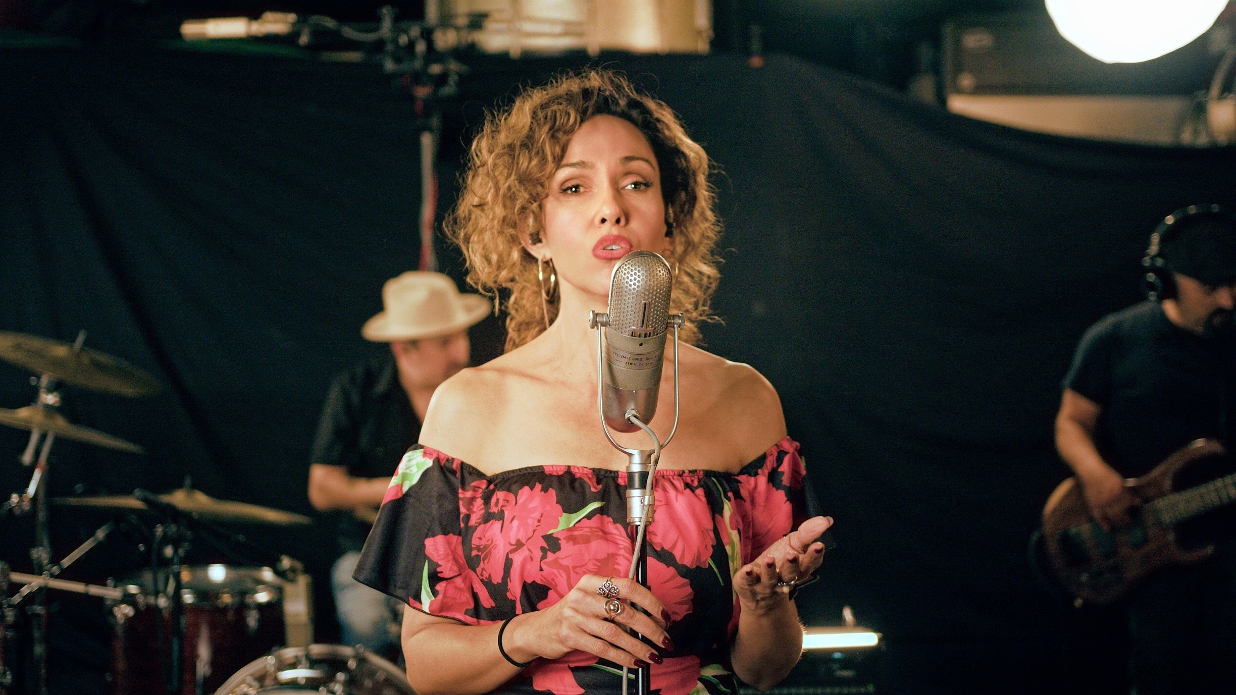 A woman sings into a vintage microphone while musicians play behind her in a dim studio.