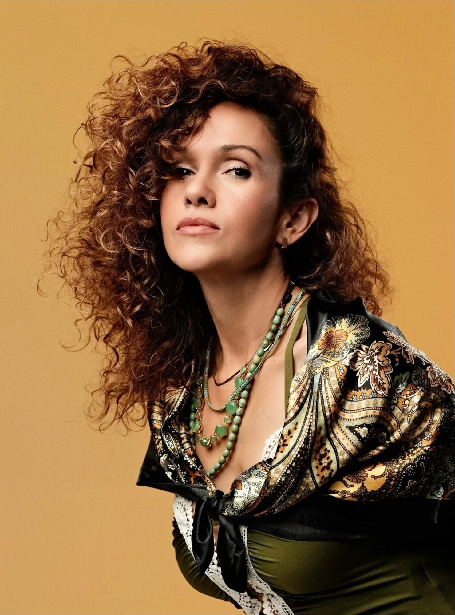 Person with curly hair wearing a patterned shawl and layered necklaces against a tan background.