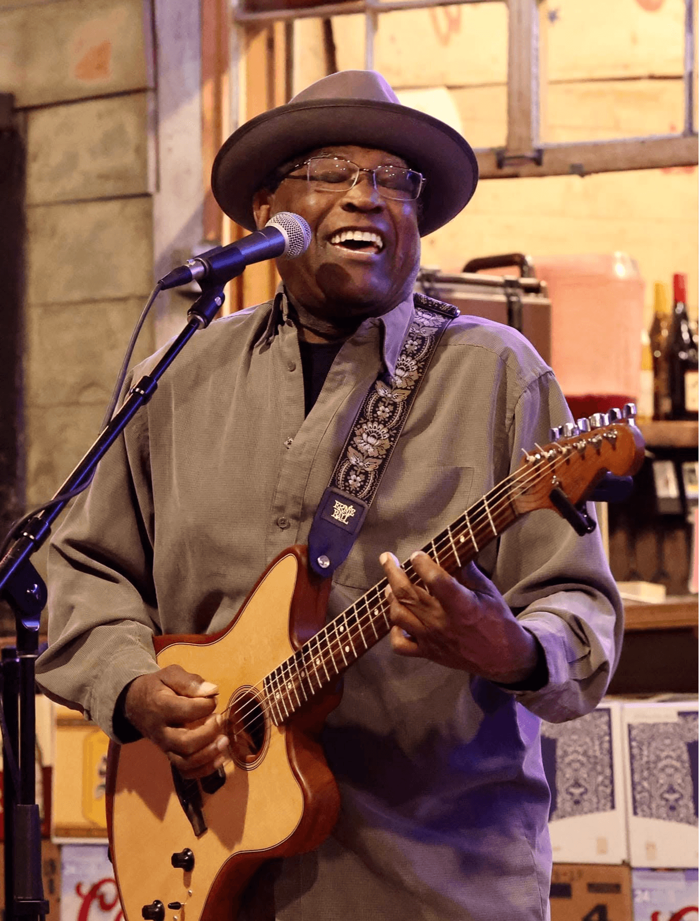 A musician wearing a hat sings into a microphone while playing an electric guitar.