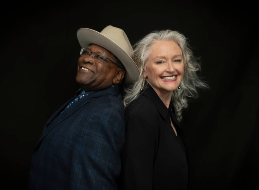 Two people pose back-to-back against a dark background; one wears a light cowboy hat and the other has gray hair.
