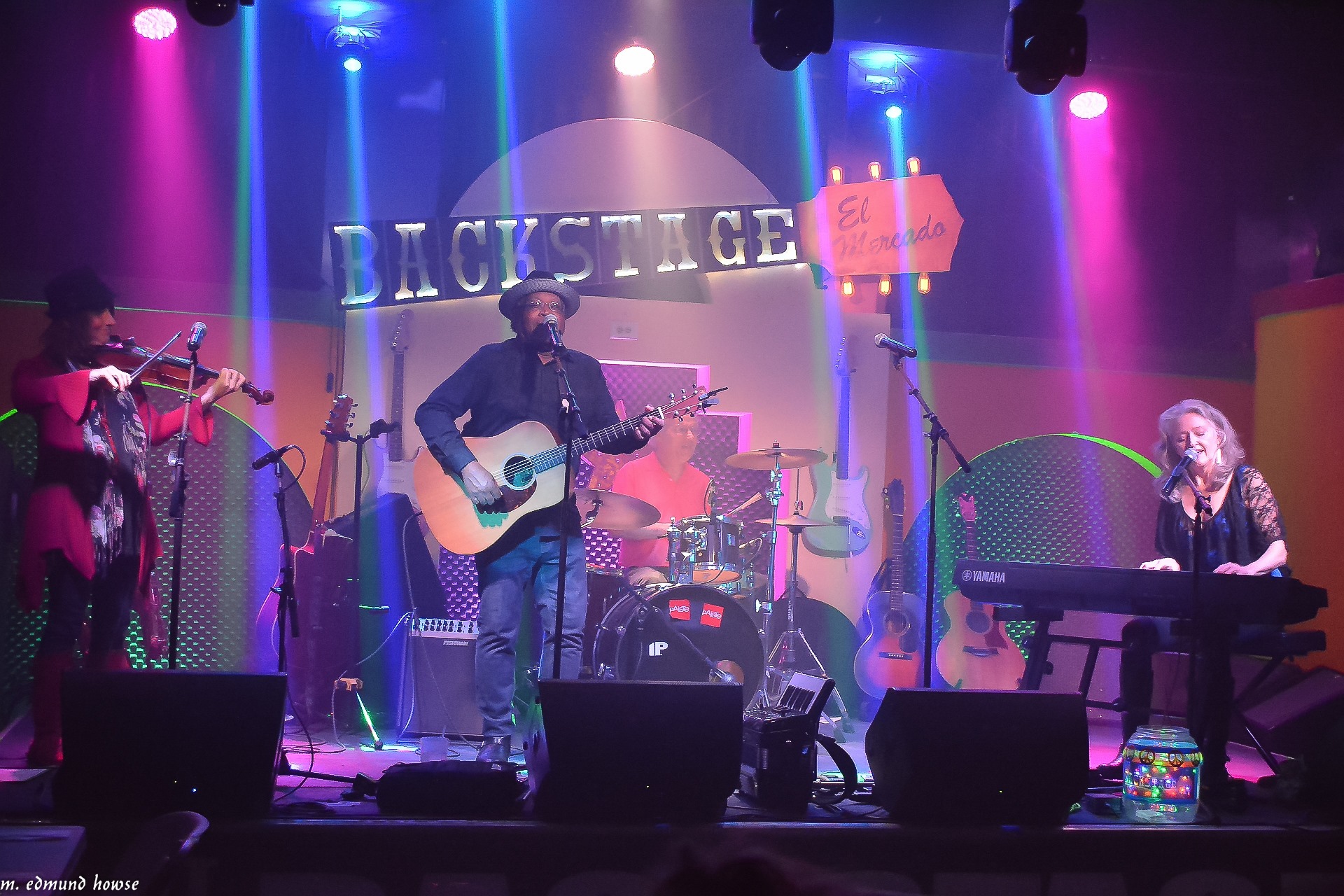 A band performs onstage under colorful lights, with a guitarist and singer, a drummer, and a keyboard player.