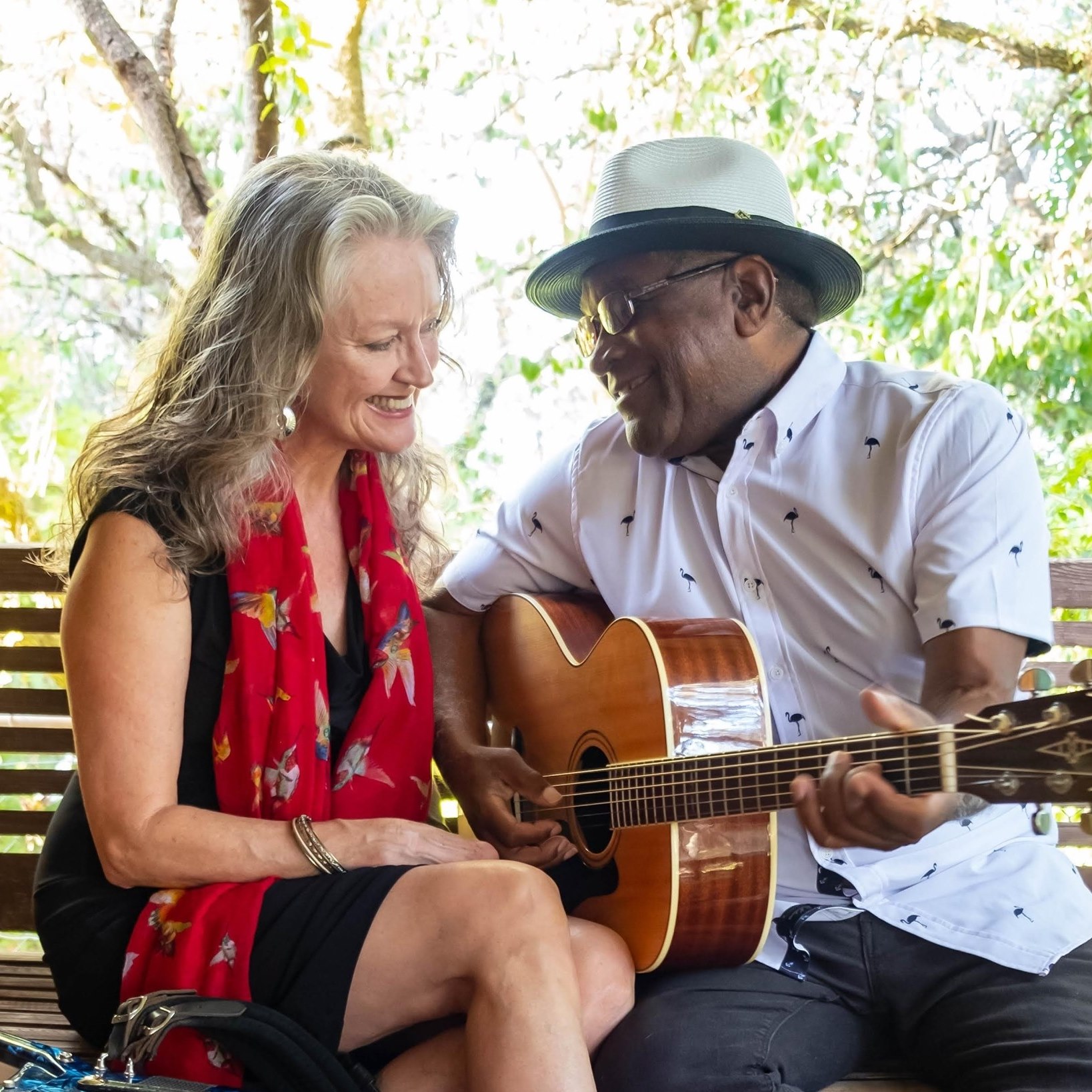 Two people sit together outdoors, smiling as one plays an acoustic guitar.