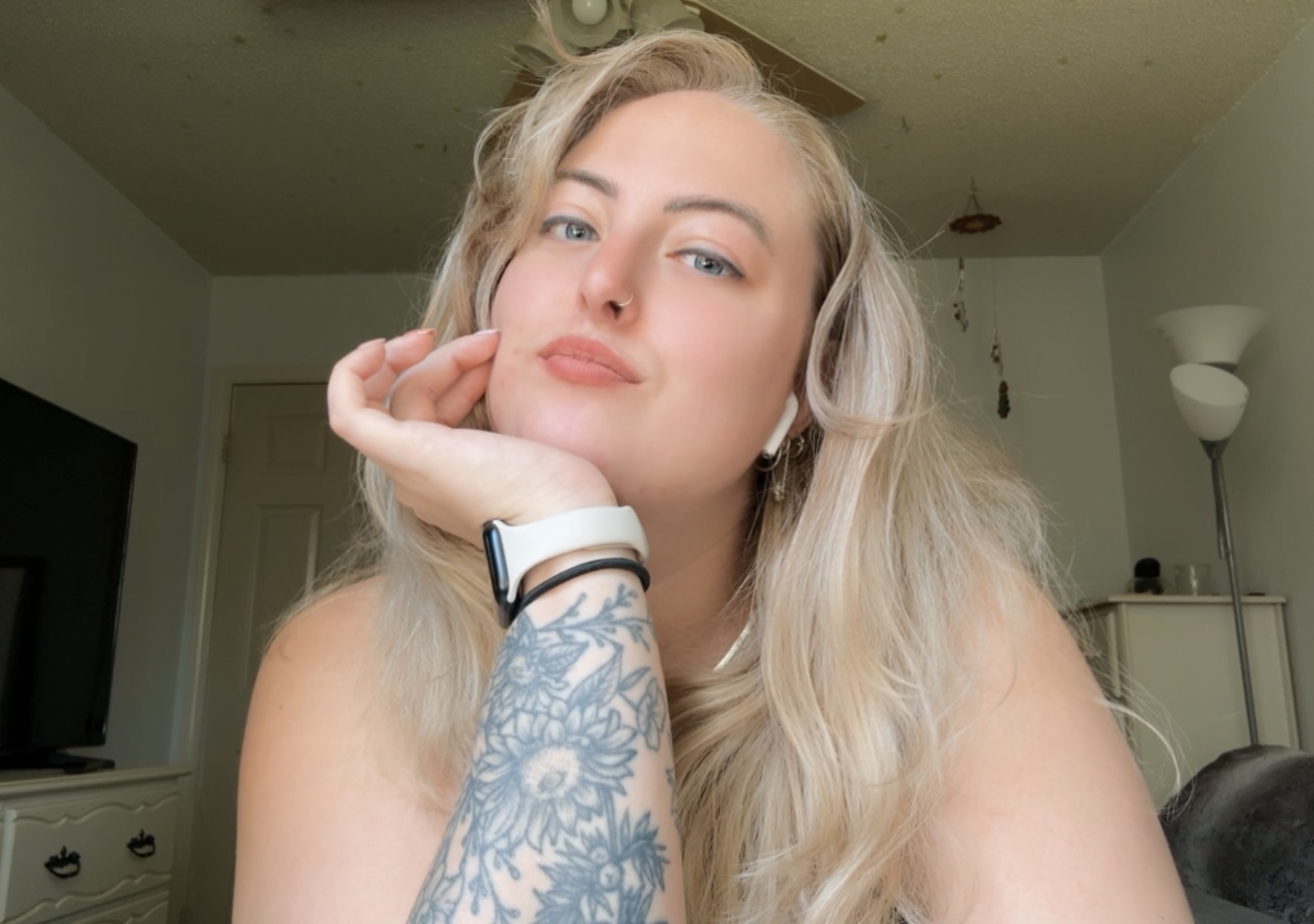 A person with long blonde hair rests their chin on a tattooed hand in a room.