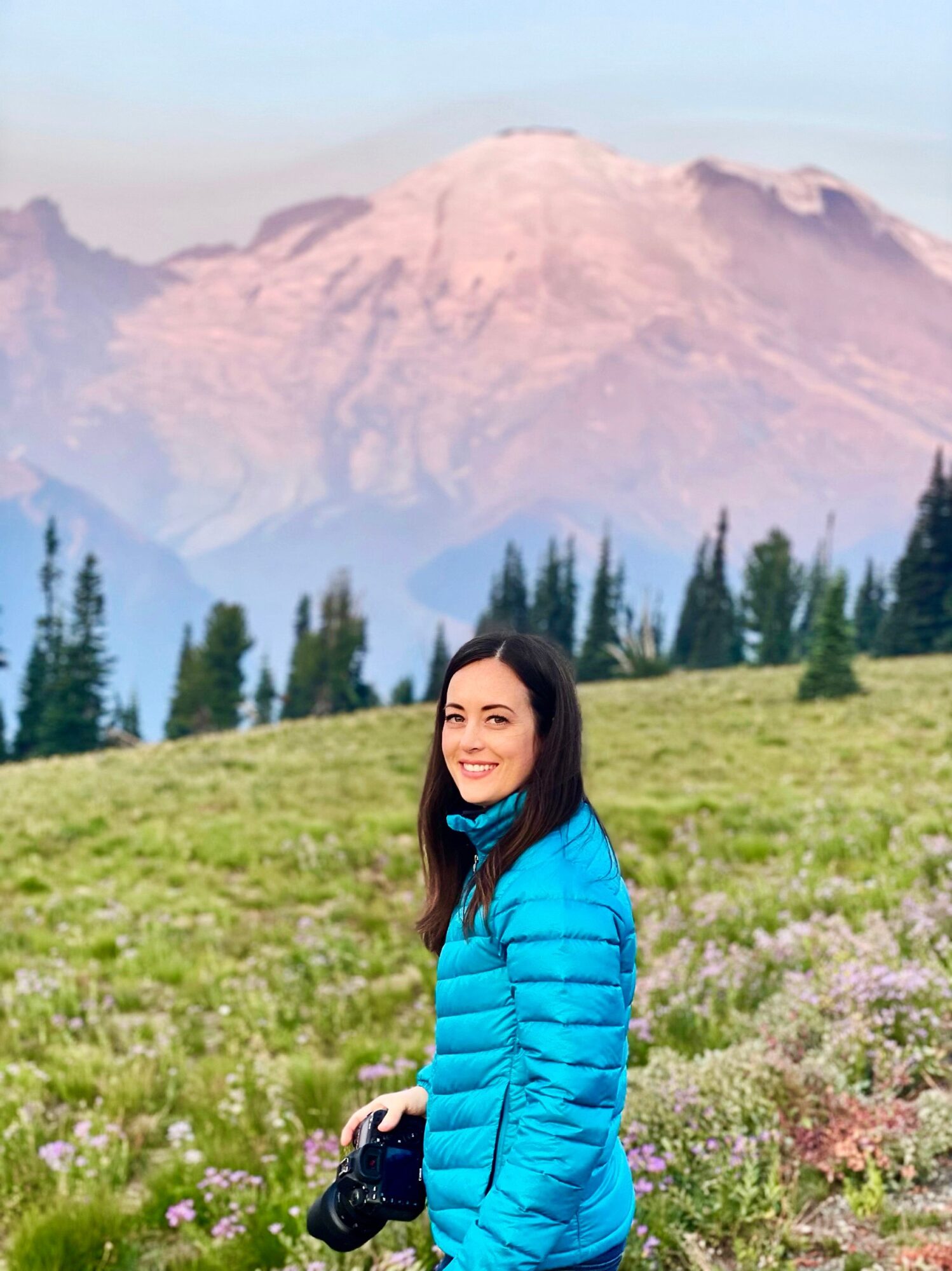 Person in a blue jacket holding a camera in a flowered meadow, with evergreen trees and a mountain behind.