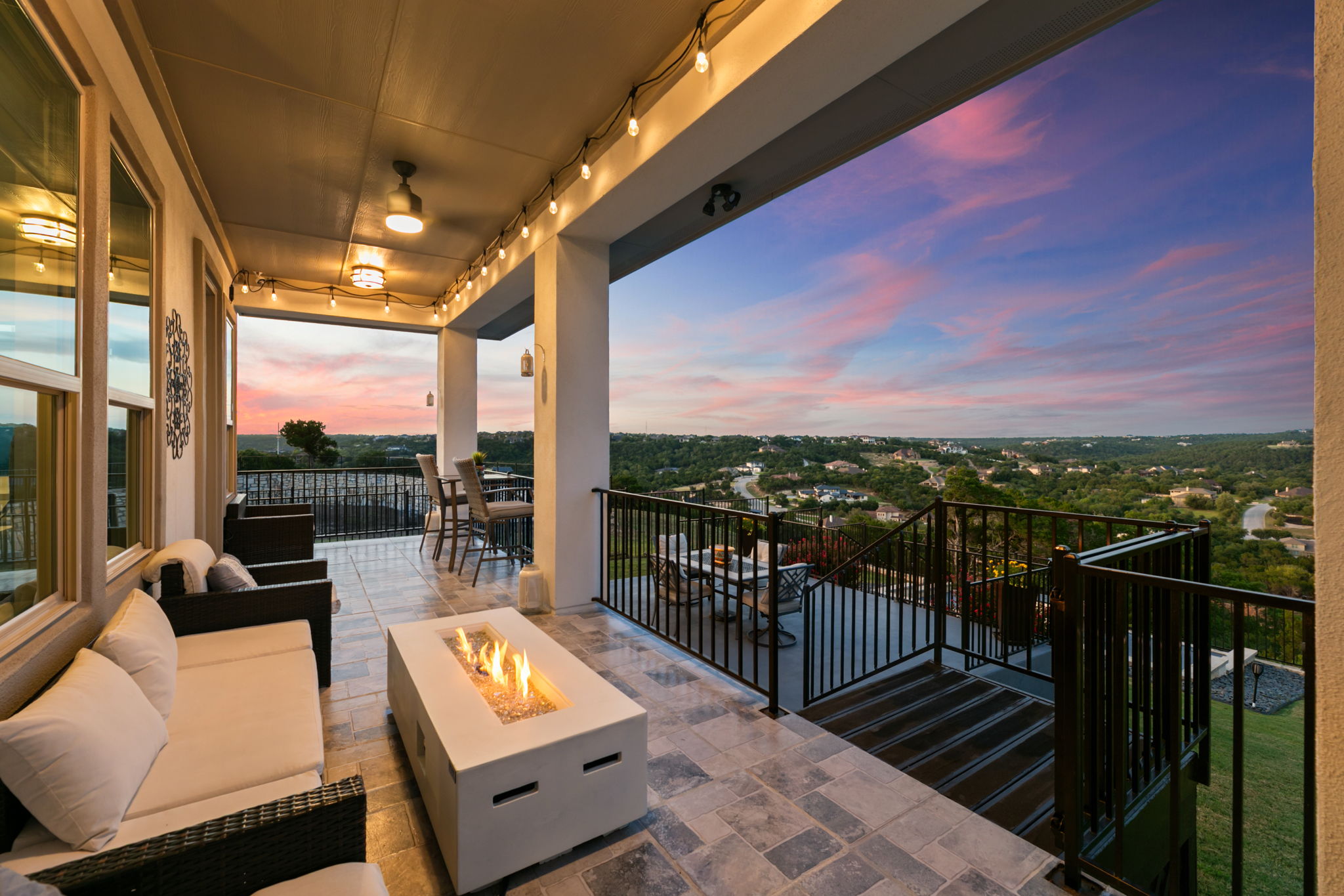 Covered patio with seating, a table, and a railing overlooking a green landscape at sunset.
