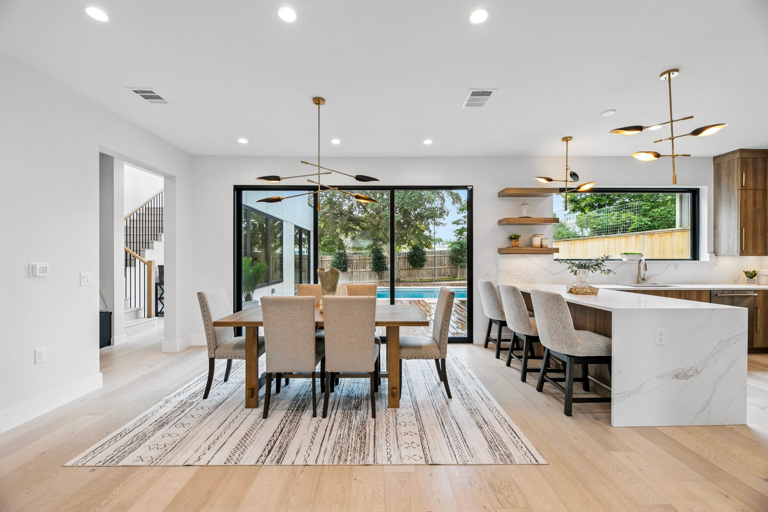 Bright open-plan dining and kitchen area with a wooden table, upholstered chairs, large windows, and a central island.