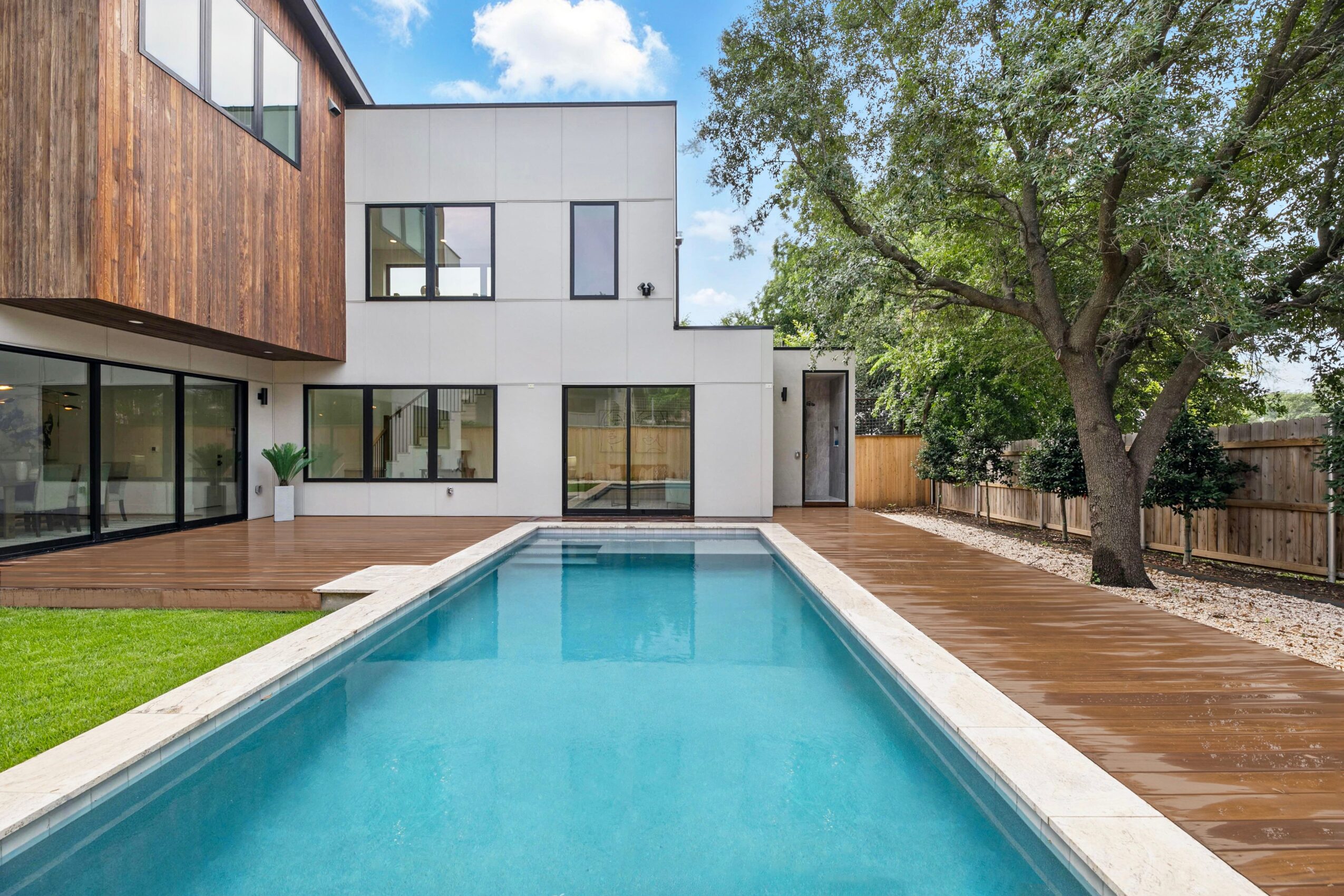 Modern two-story house beside a rectangular swimming pool, wooden deck, lawn, and large tree.