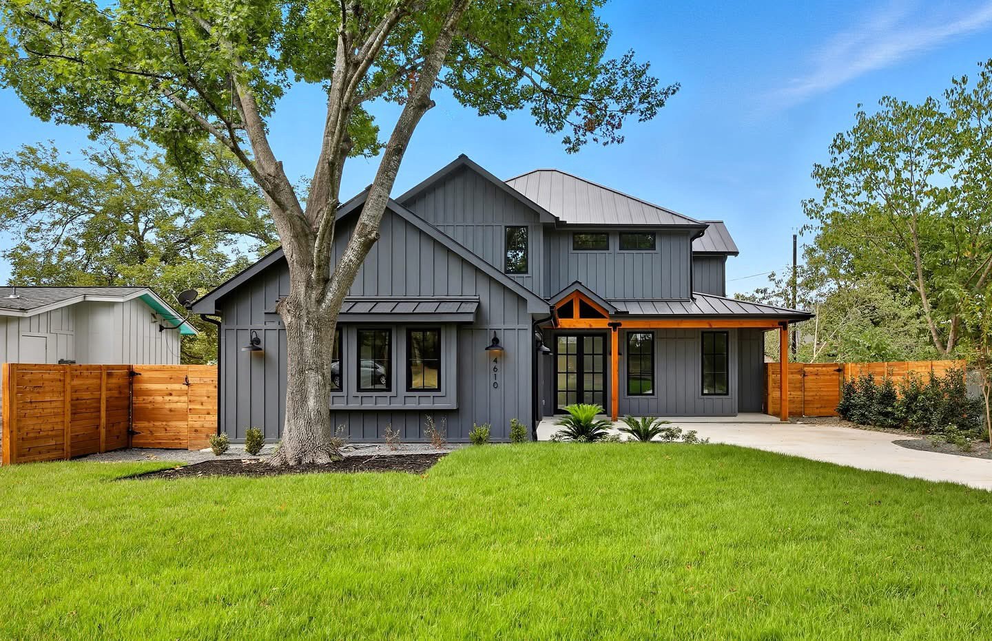 Gray modern house with a large tree, wooden fences, driveway, and green lawn beneath a blue sky.