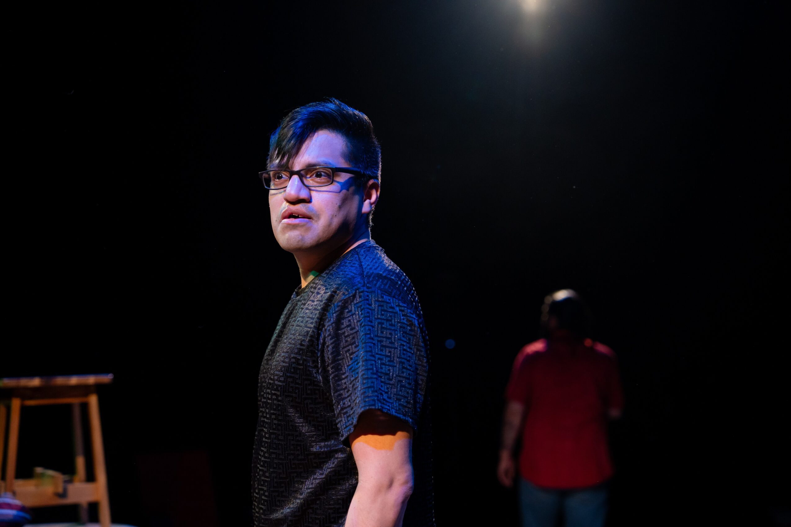 A person in glasses stands under a spotlight while another person in a red shirt walks in the background.